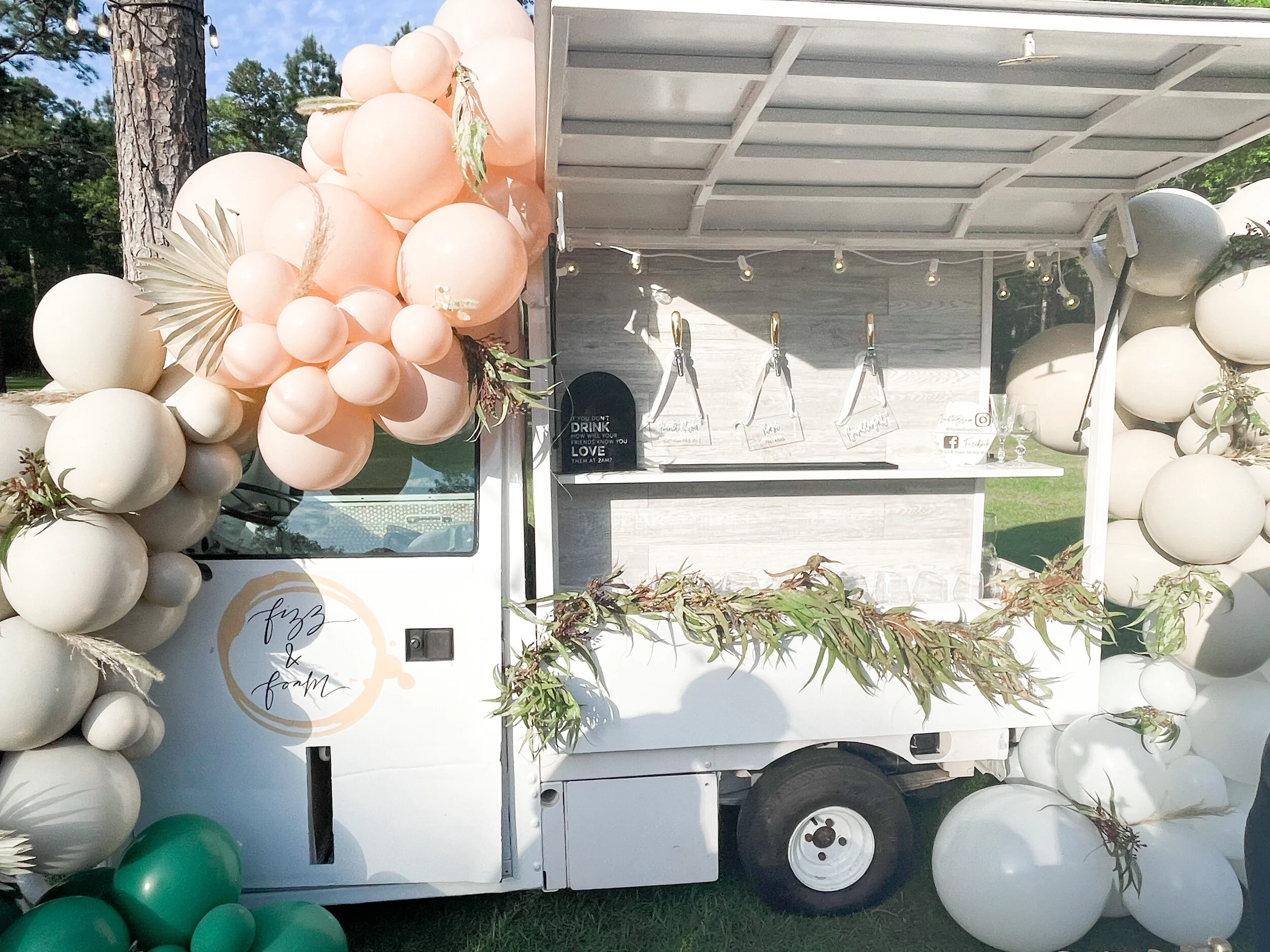White food truck decorated with pink, white, and green balloons, and greenery, set outdoors at a sunny event.