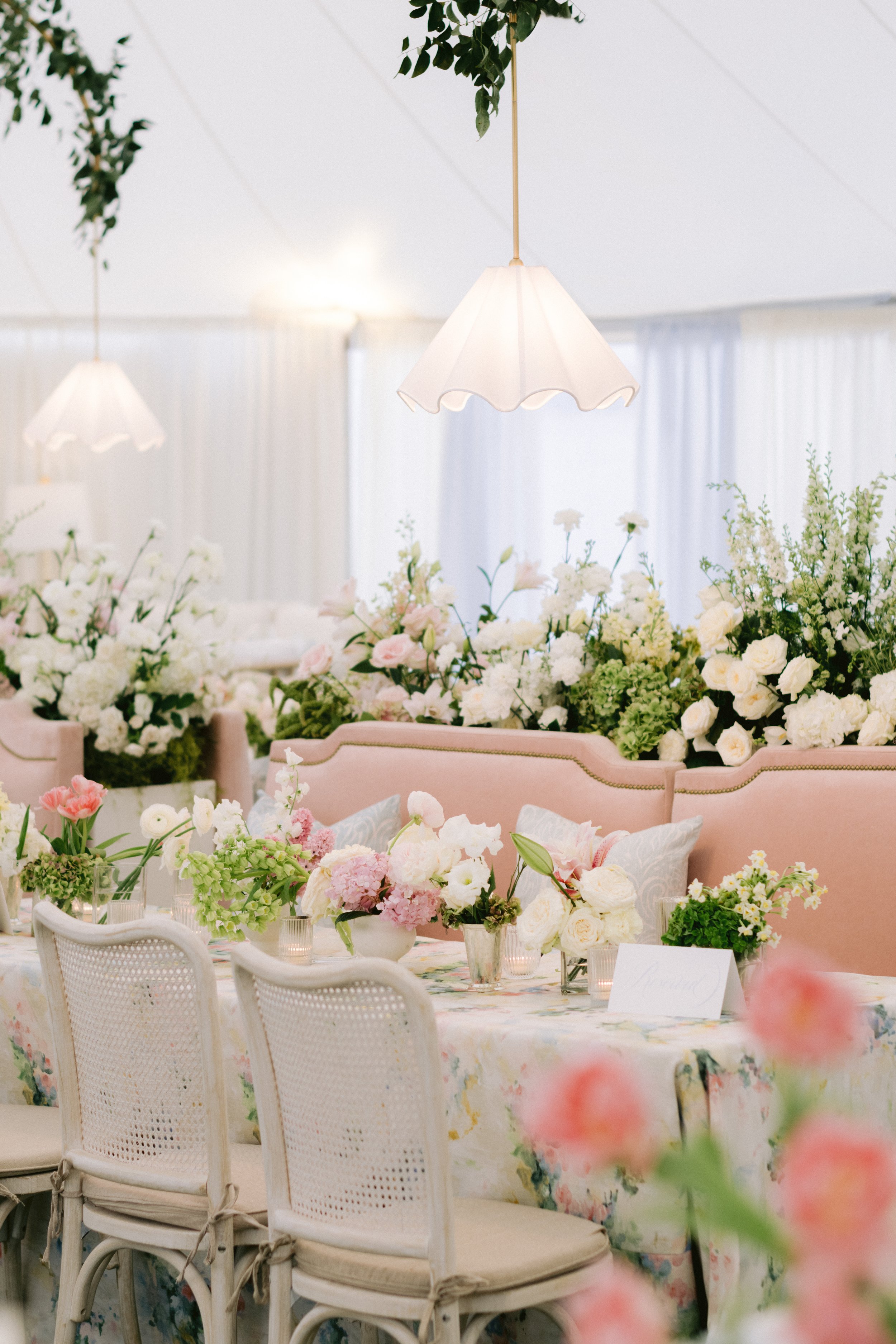 Elegant indoor event setting with floral arrangements, white chairs, and a table with a floral tablecloth, featuring soft lighting and hanging lamps.