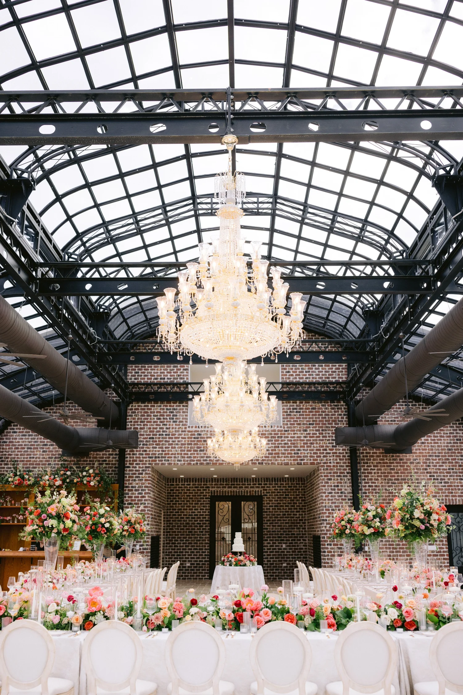 Completed reception installation with a long decorated guest table, white chairs and overhead crystal chandeliers