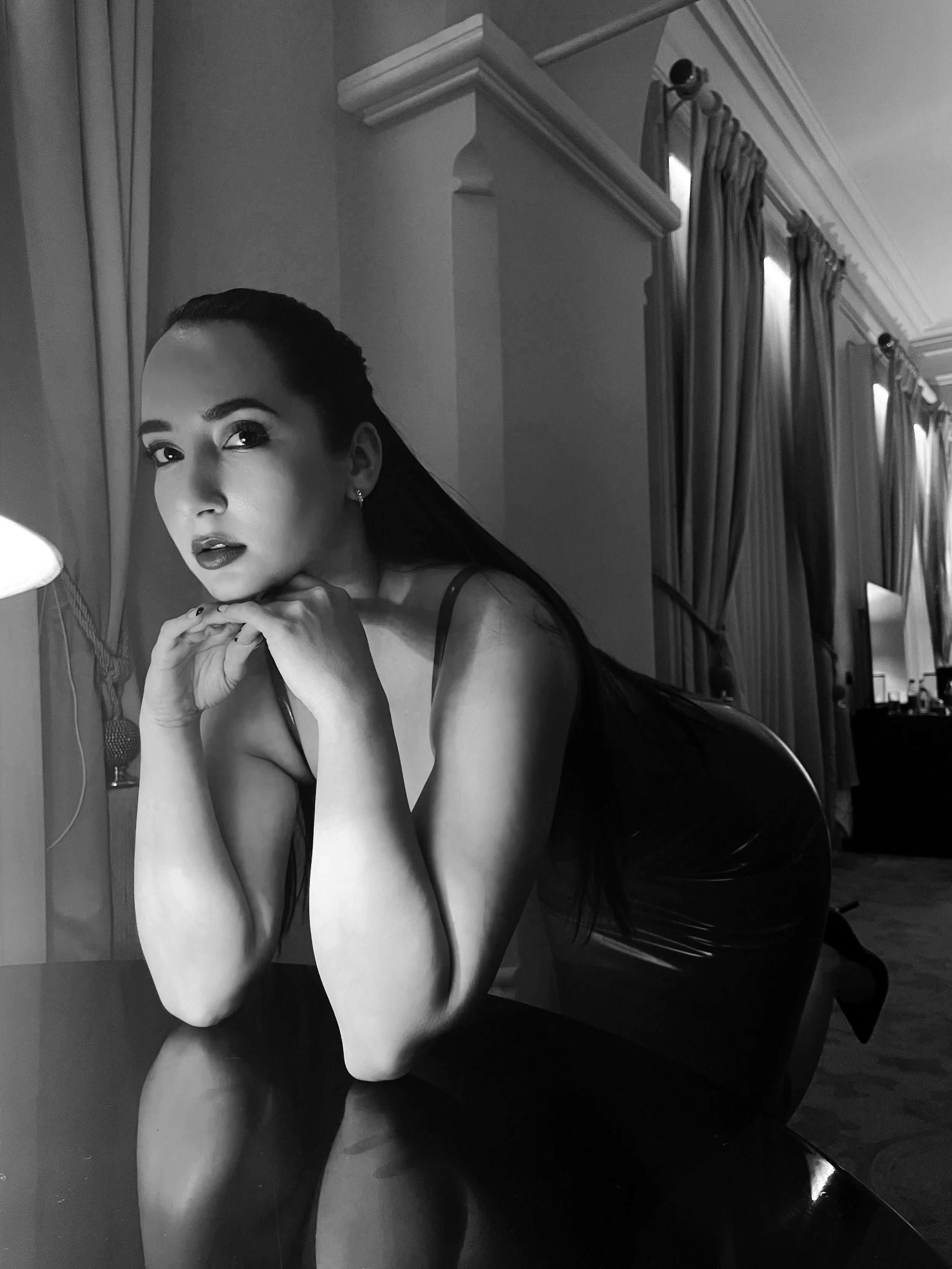 A black and white photo of a woman with long dark hair, wearing a sleeveless dress, sitting at a table with her chin resting on her hands and looking at the camera in a decorated room with curtains and windows.