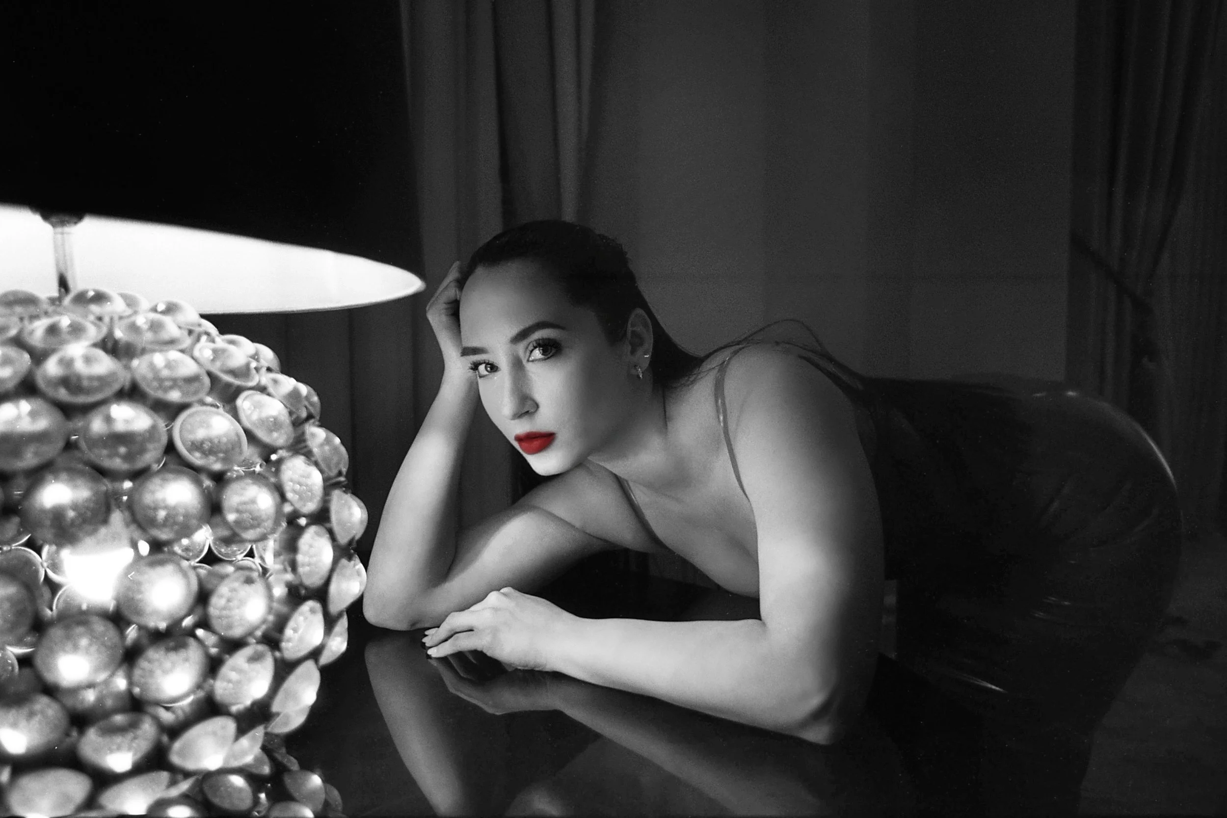 Black and white photograph of a woman with dark hair, wearing a sleeveless top with spaghetti straps, lying on her stomach on a reflective surface. She rests her head on her hand and looks at the camera with a serious expression. A large, shiny, deco