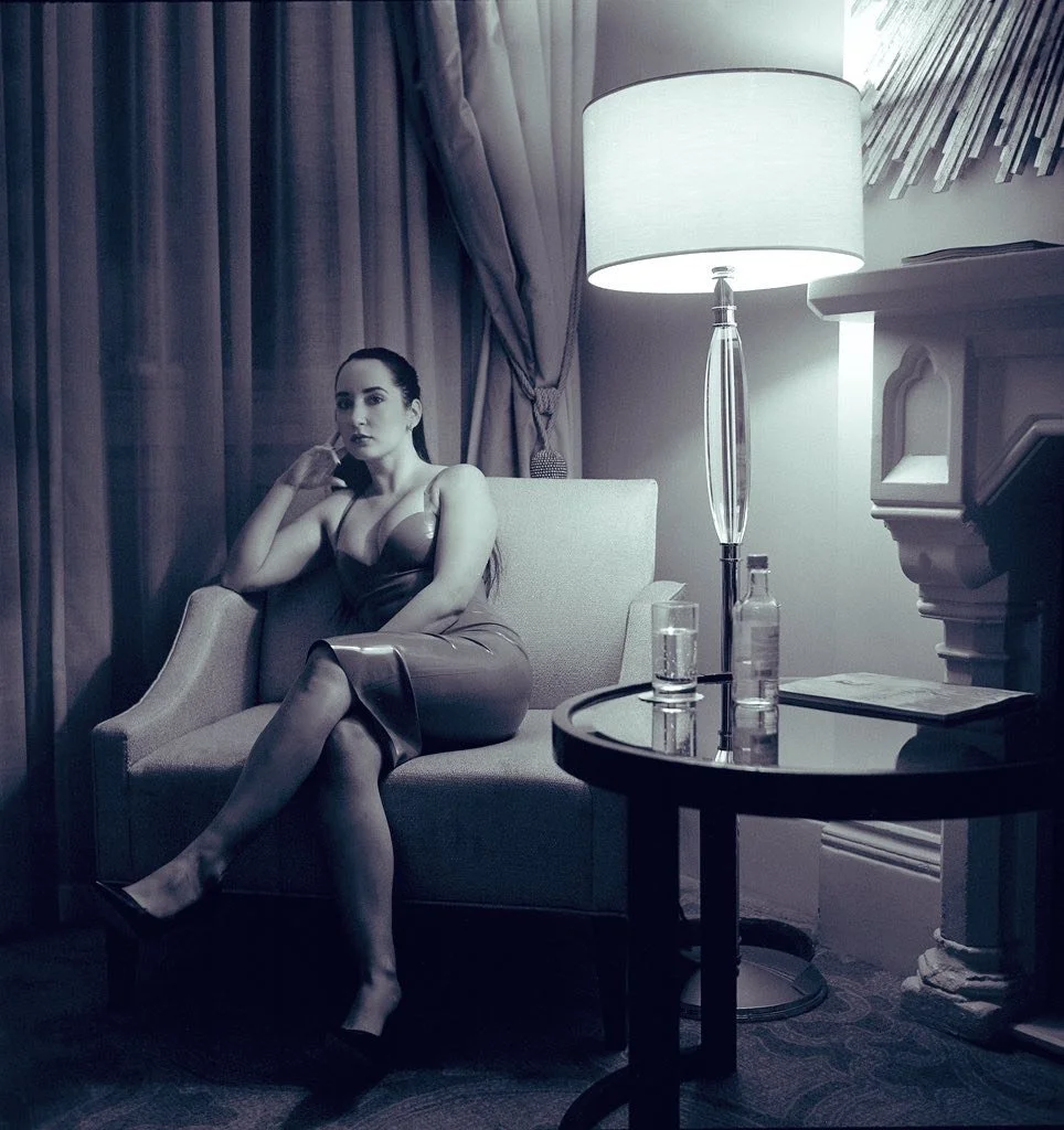 A woman sitting on a beige armchair in a dimly lit room, wearing a dress, with a side table holding a glass of water, a bottle, and a magazine, near a lamp and curtains.