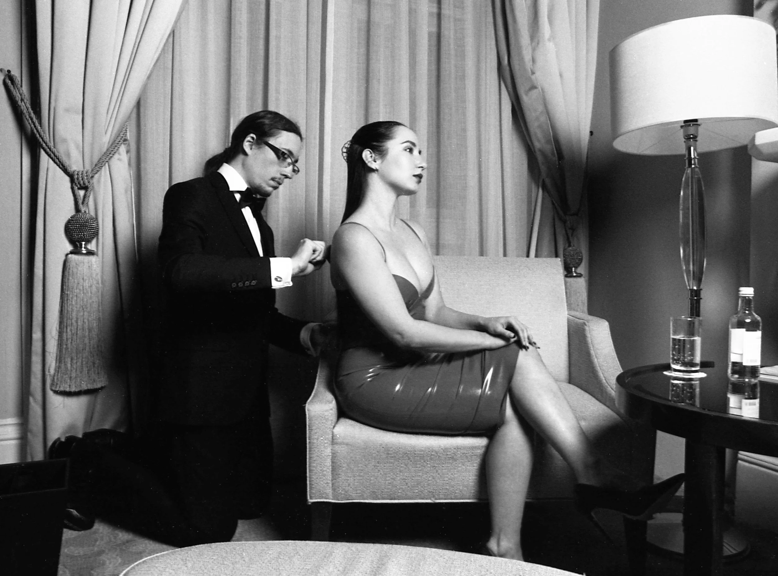 A woman is sitting on a sofa while a man adjusts her dress, in a room with curtains and a table with a bottle and a glass.