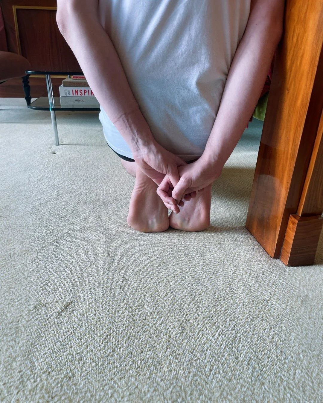 Person kneeling on beige carpet with their hands clasped and head bowed, next to a wooden piece of furniture, with a glass table and books in the background.