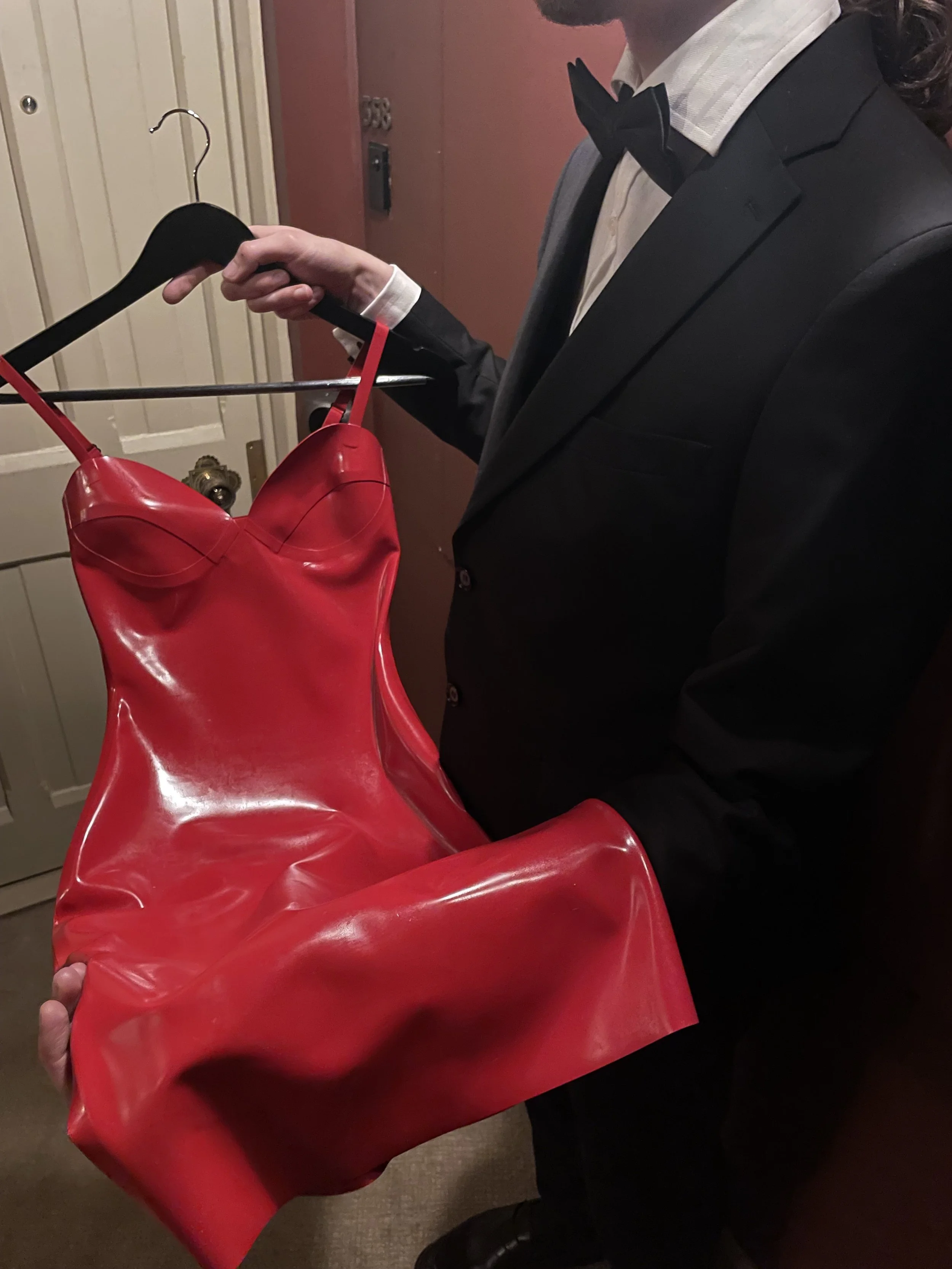 Person in a tuxedo holding a red, shiny, leather dress that resembles a form-fitting, stylized dress.