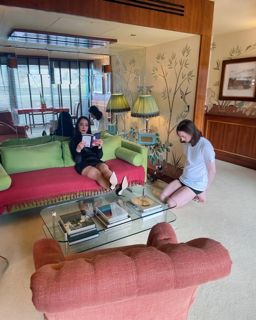 A woman in black dress and stockings sits on a green couch reading a book, while another woman in a white shirt kneels on the floor near her, looking down. A third woman in white shirt and black shorts sits in the background at a dining table. The ro