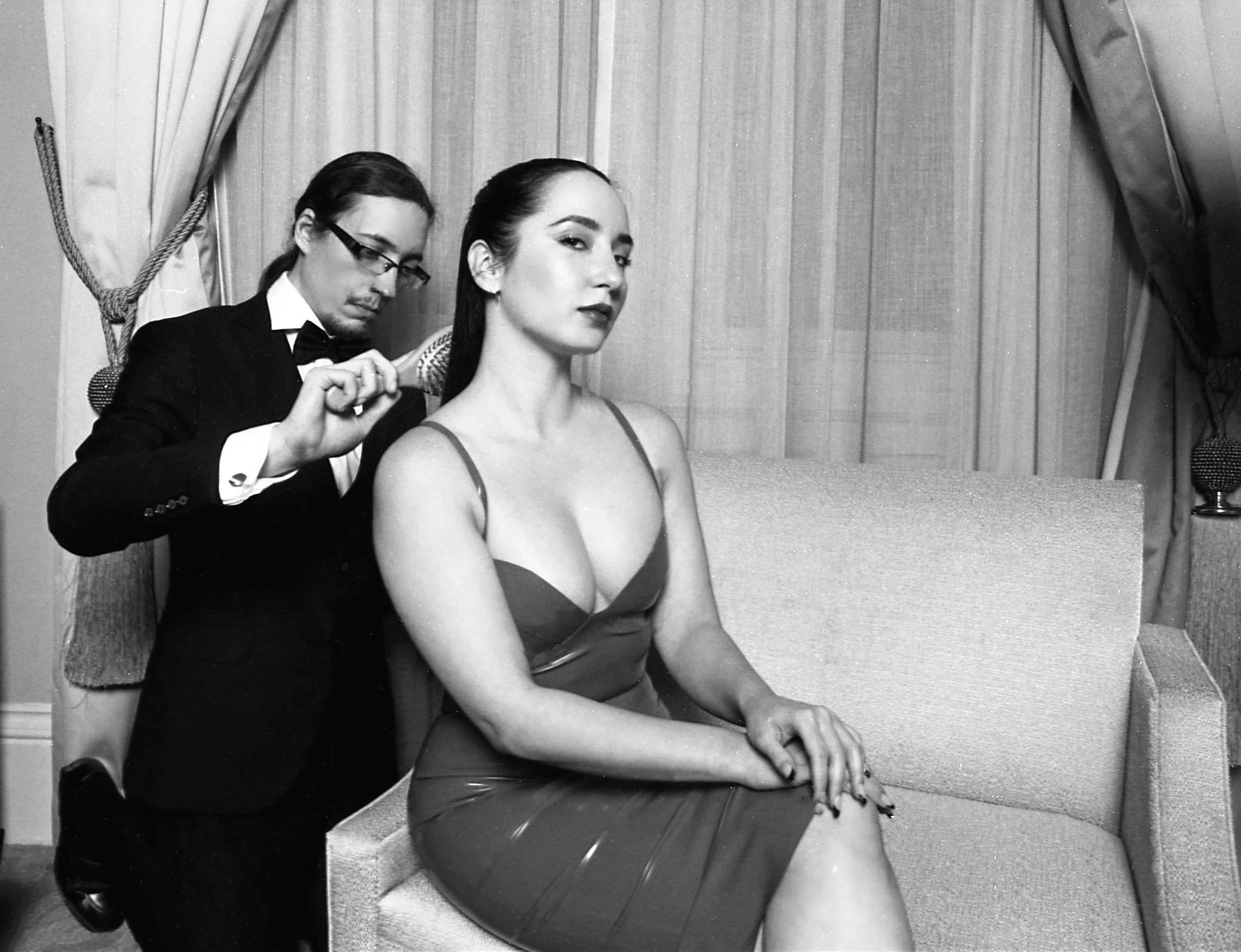 A woman with dark hair and striking makeup sitting on a couch with her hands on her lap, while a man in a tuxedo is combing her hair behind her in a room with curtains.