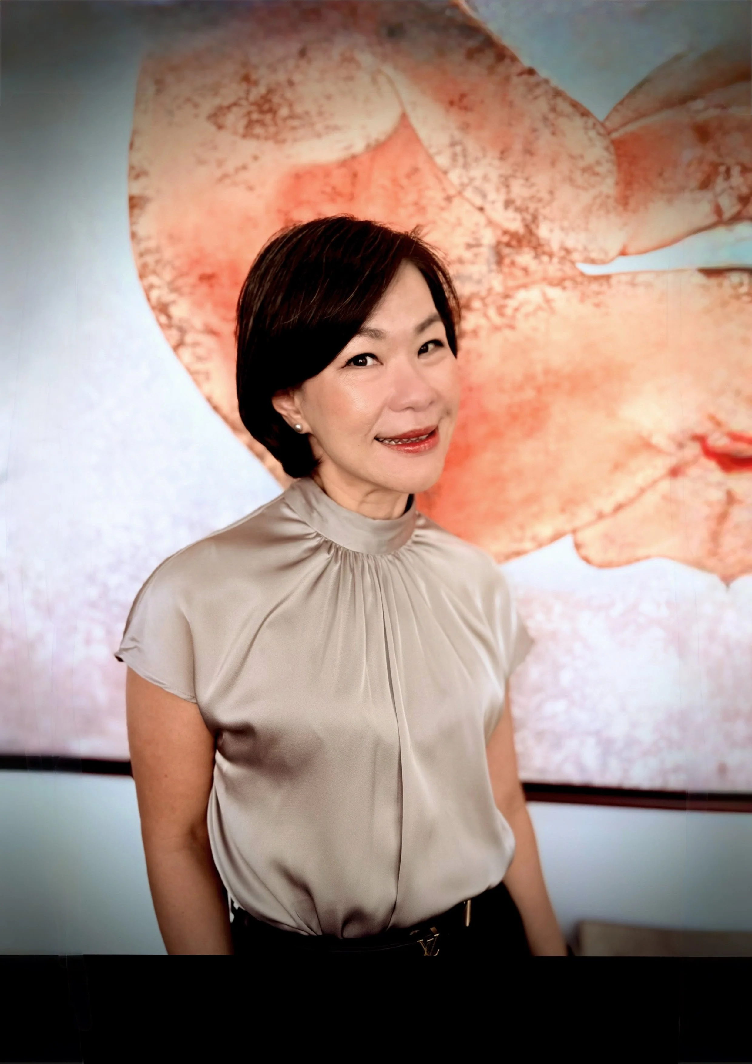 Keny Wardhana & Co with short dark hair, wearing a shiny beige blouse, standing in front of a large, colorful abstract artwork.