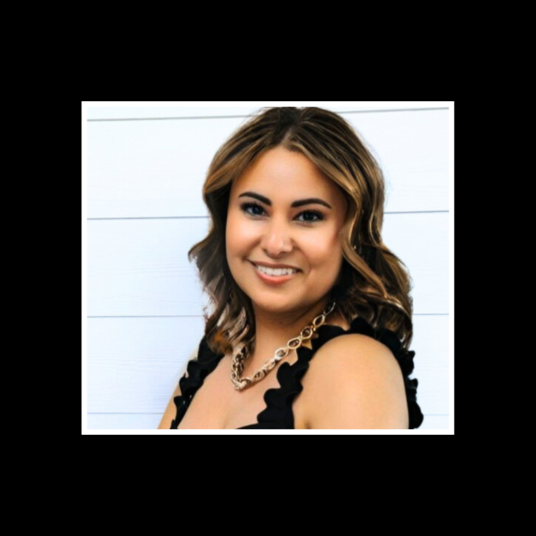 A woman with light brown, wavy hair wearing a black top with ruffled straps and a gold chain necklace, smiling in front of a white wooden wall.