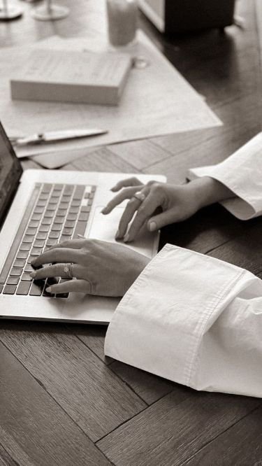 Person typing on a laptop keyboard at a desk with a notebook, pen, paper, and a cup in the background.