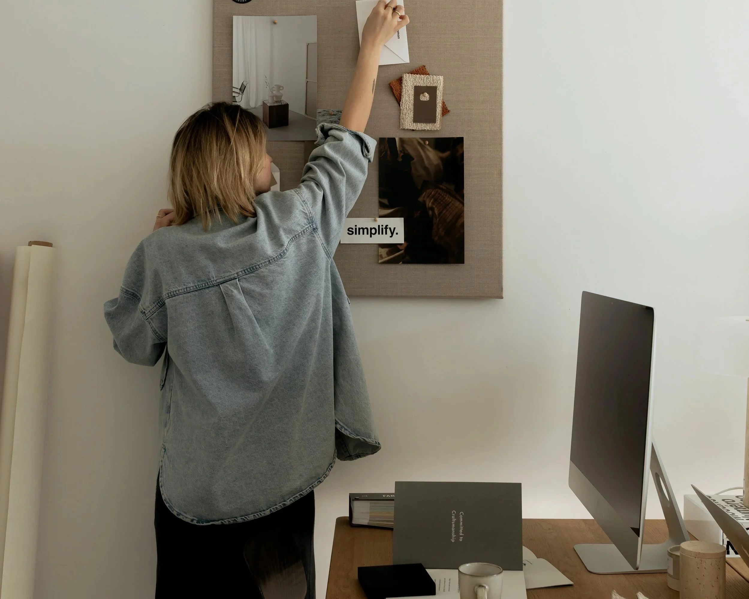 A woman with blonde hair wearing a denim jacket is standing in front of a beige display board, reaching up to place or adjust something on the board. The display board has various items attached, including a mirror, a photo, and a small artwork. There are books, cups, and a computer monitor on the desk below the display. The setting appears to be an office or a creative workspace.