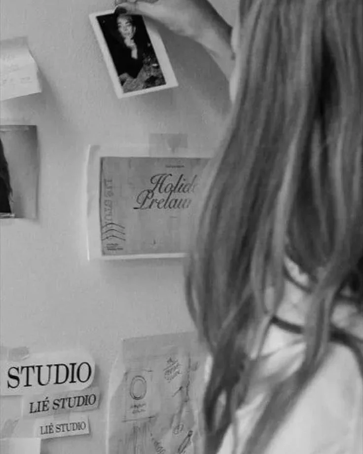 A person with long hair and glasses is standing next to a wall with papers and pictures, including a sign that says "STUDIO" and a framed certificate or document.