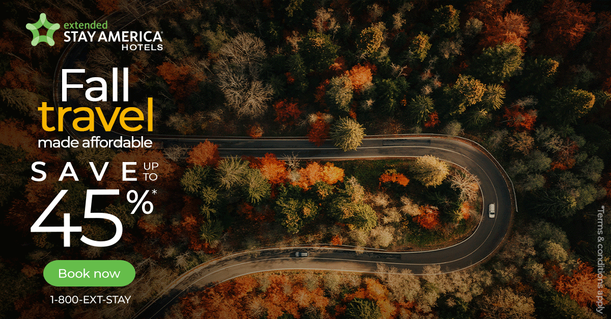 Aerial view of a winding road through a forest with colorful fall foliage, overlaid with promotional text for Stay America Hotels offering up to 45% discount, with a green booking button and contact number.