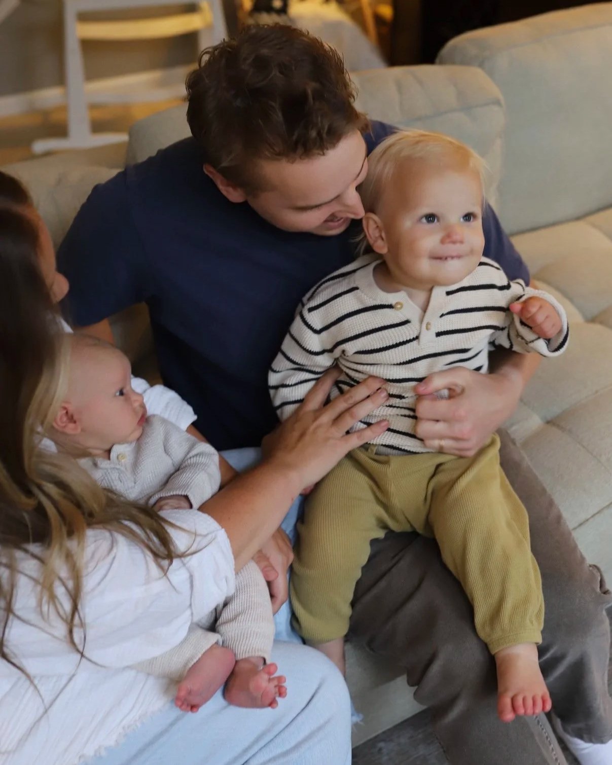 A man sitting on a couch holding a young child in his lap, smiling and looking at him. Two young girls are nearby, one touching the child's arm and the other sitting with her legs crossed. The scene appears to be in a cozy, indoor setting.