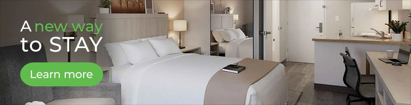 Hotel room interior with a bed, nightstands, lamps, a sitting area, and a kitchenette. Overlay text: 'A new way to STAY' with a green button saying 'Learn more.'