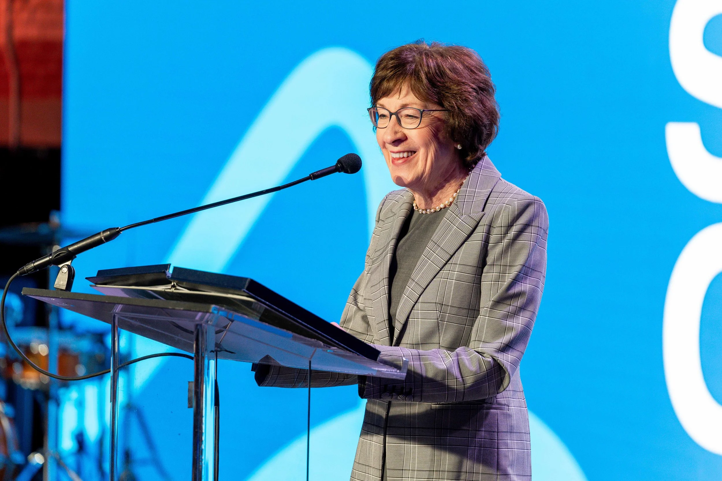 A woman with short brown hair and glasses, wearing a gray blazer and pearl necklace, standing at a podium giving a speech at an event with a large blue background.