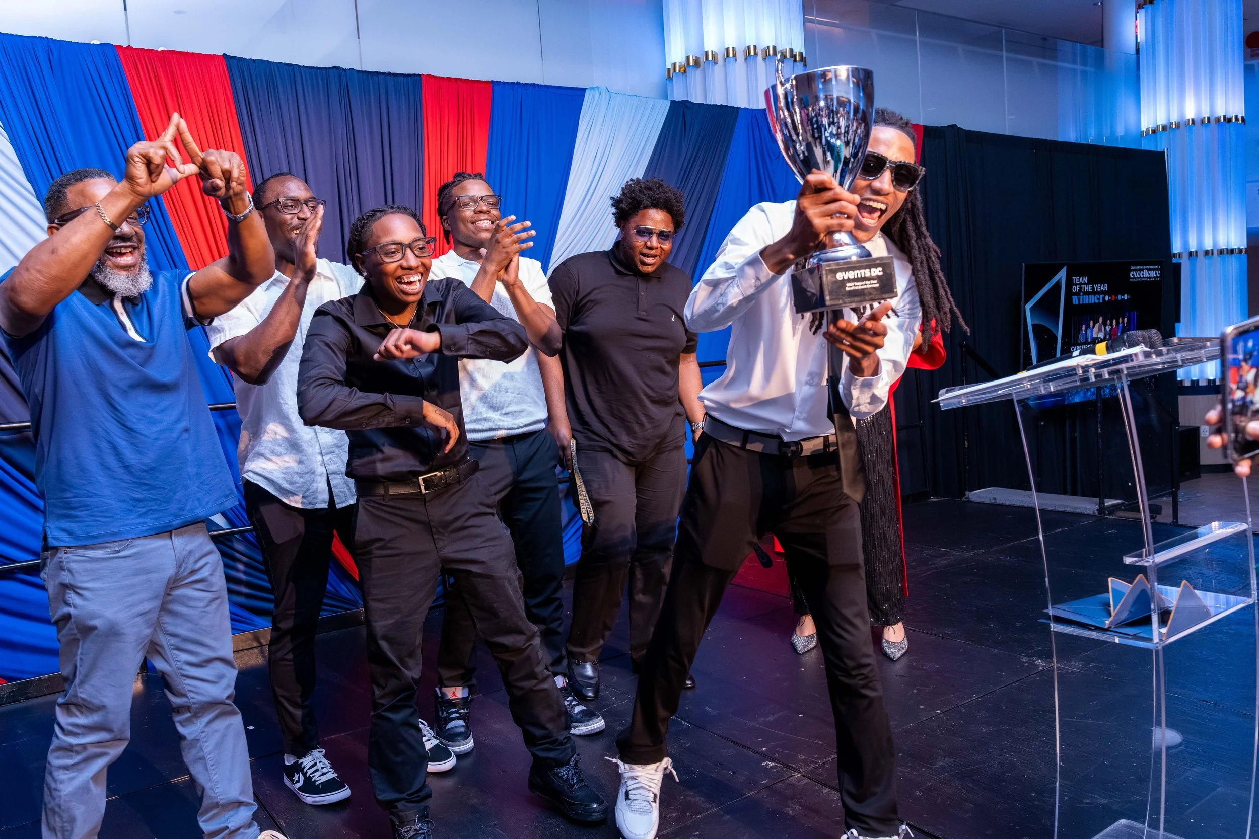 A group of people celebrating on stage at an awards ceremony, with one person holding a trophy and wearing sunglasses, while others clap and cheer.