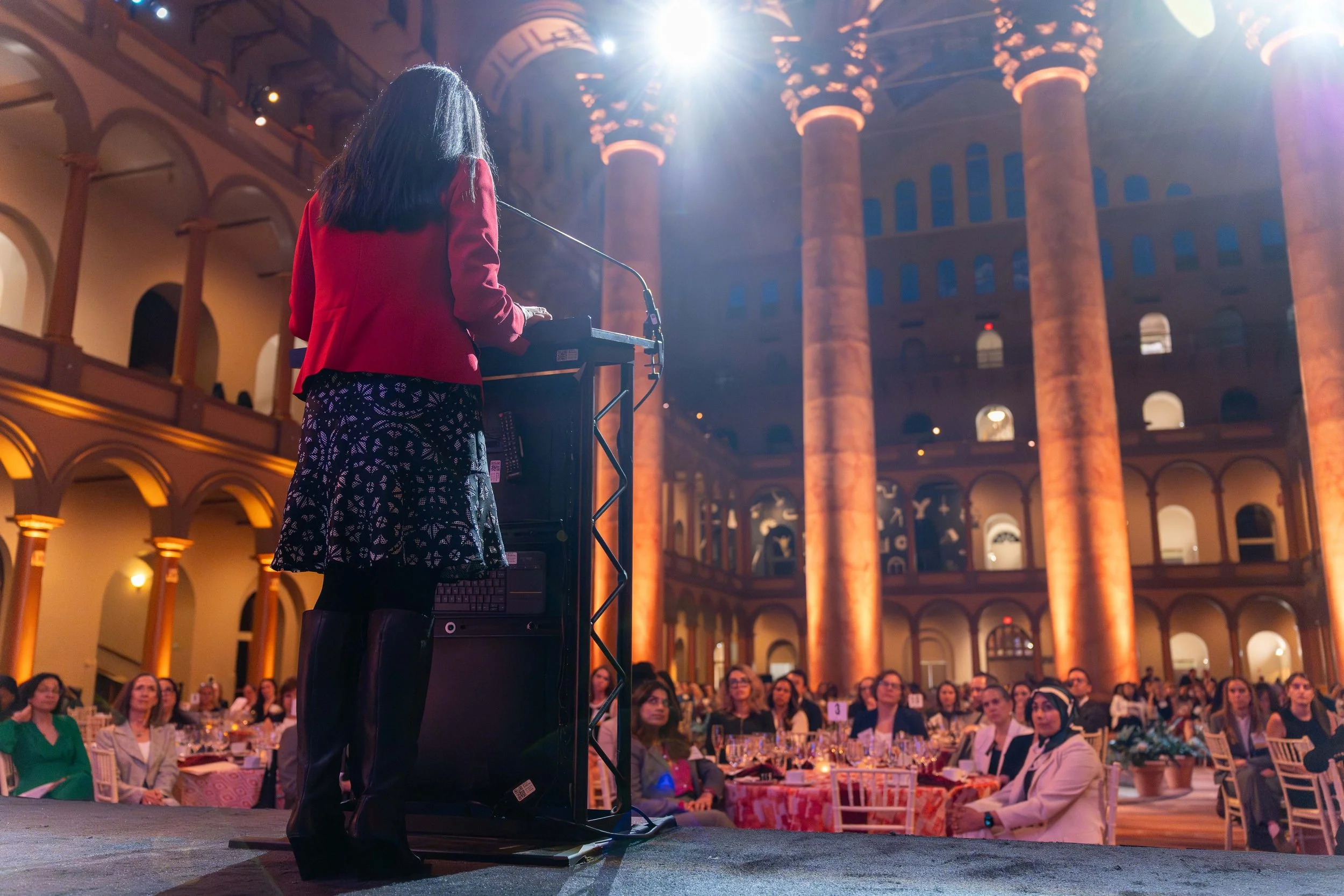 A woman in a red jacket and patterned skirt speaking at a podium at the National Building Museum, facing an audience seated at tables during a formal event.