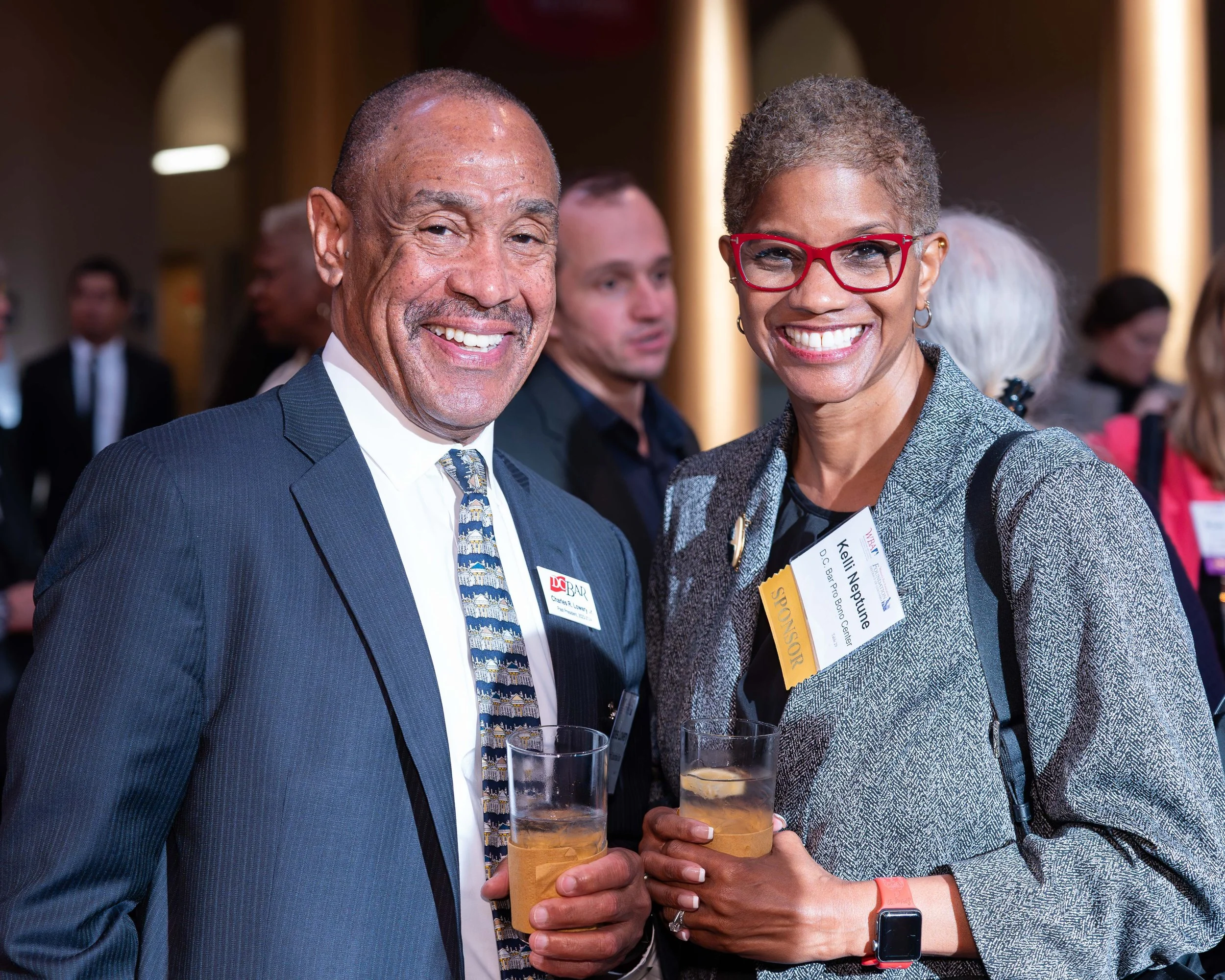 Two smiling professionals at a networking event holding drinks, one man in a suit and a woman with glasses and a gray blazer.