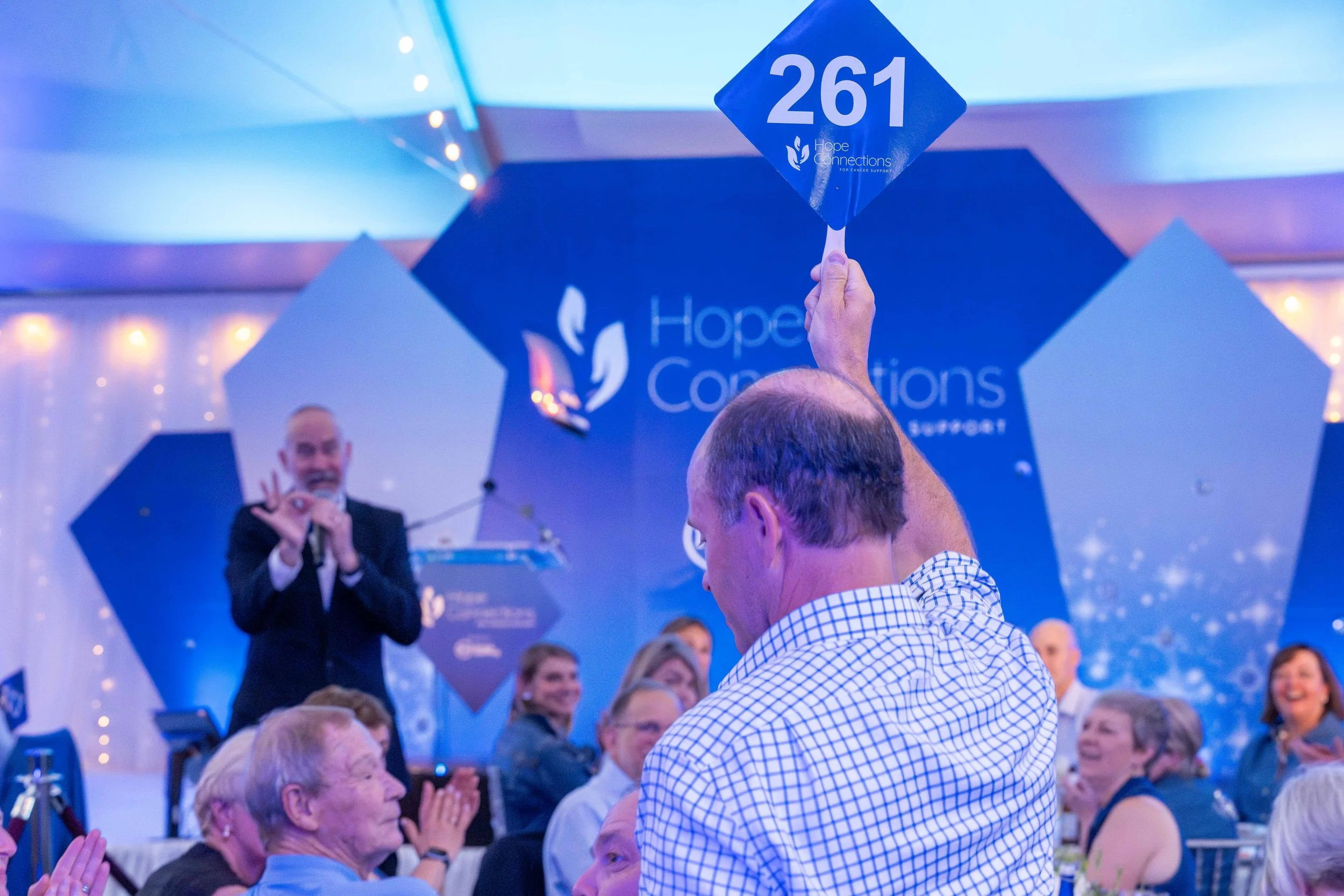 A man is holding a blue sign with the number 261 at a Hope Connections gala, with a speaker and seated attendees visible in the background.