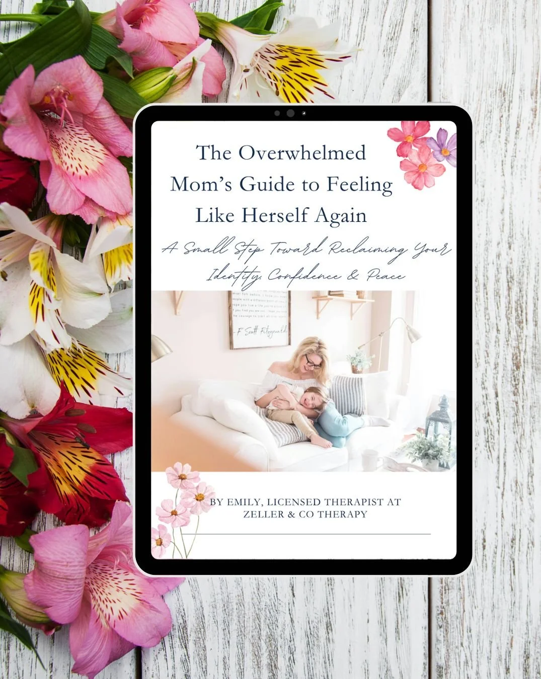 Book cover titled "The Overwhelmed Mom's Guide to Feeling Like Herself Again" with a subtitle "A Small Step Toward Reclaiming Your Identity, Confidence & Peace," by Emily, licensed therapist at Zeller & Co Therapy. The cover features a cozy living room scene with a mother and child smiling on a white couch, surrounded by pink, white, and purple flowers, with a light and airy room in the background.