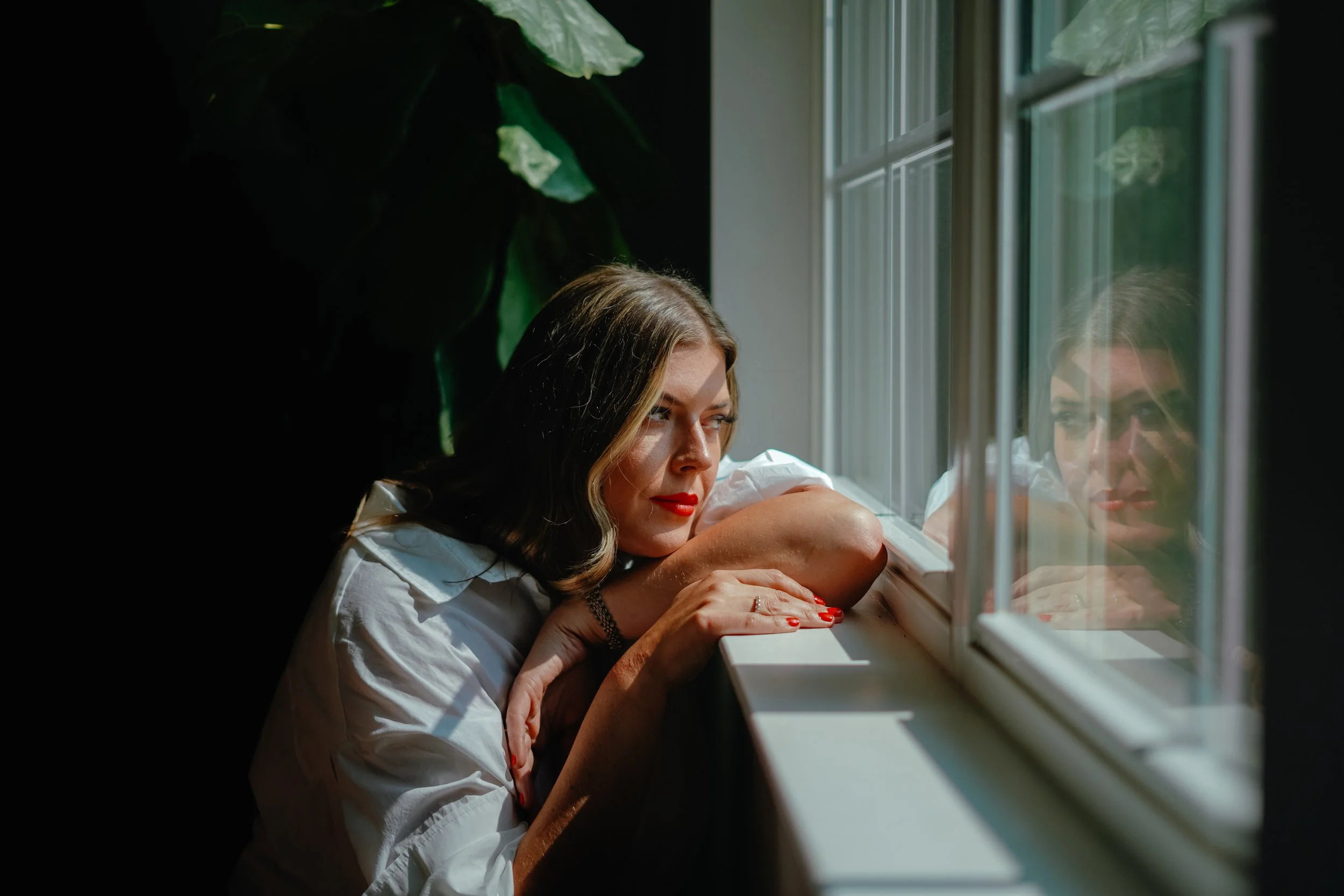 A woman with shoulder-length dark hair and red lipstick sits by a window, resting her head on her crossed arms on the windowsill, and gazes thoughtfully outside. Her reflection can be seen in the window, and sunlight illuminates her face.