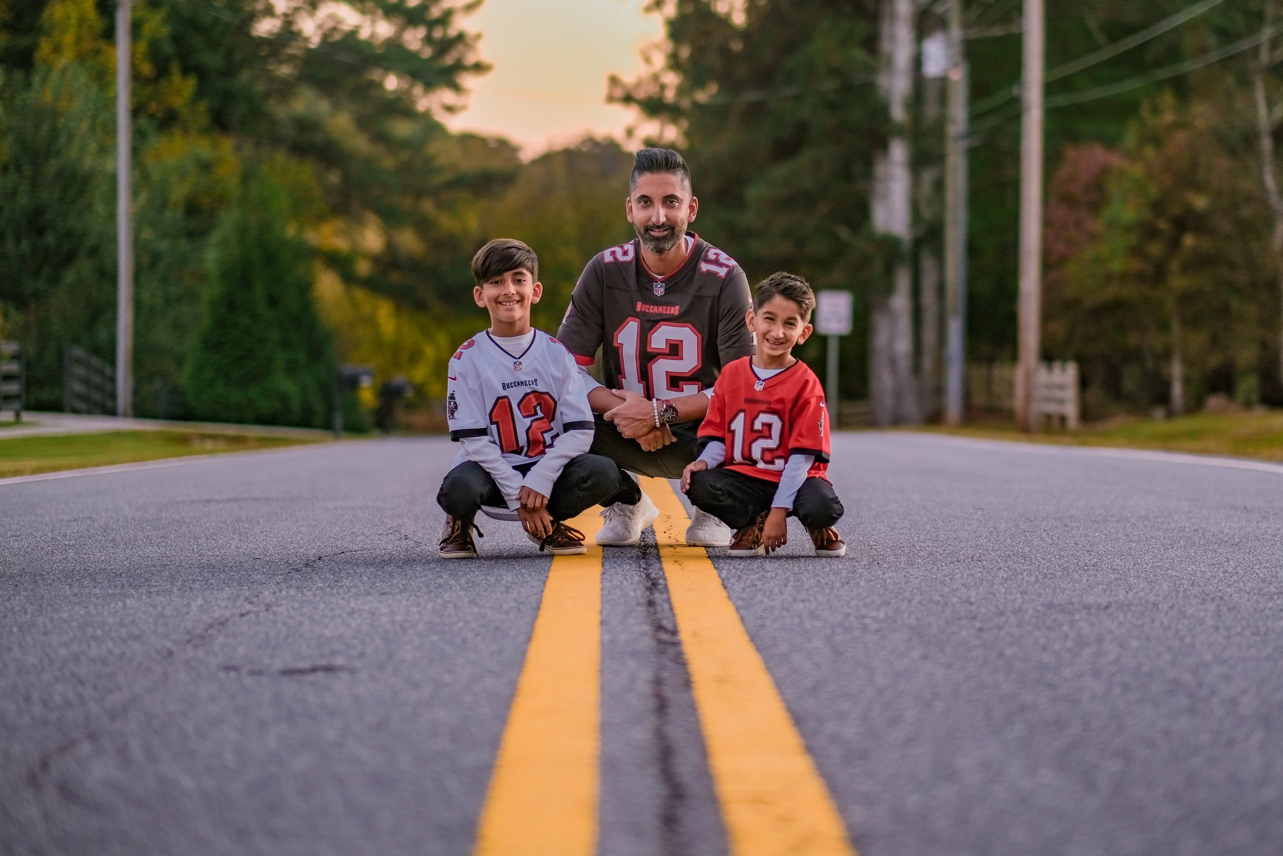 A man and two boys wearing Tampa Bay Buccaneers football jerseys with the number 12, squatting on a road with a double yellow line, shaking hands and smiling. Sunset with trees and utility poles in the background.