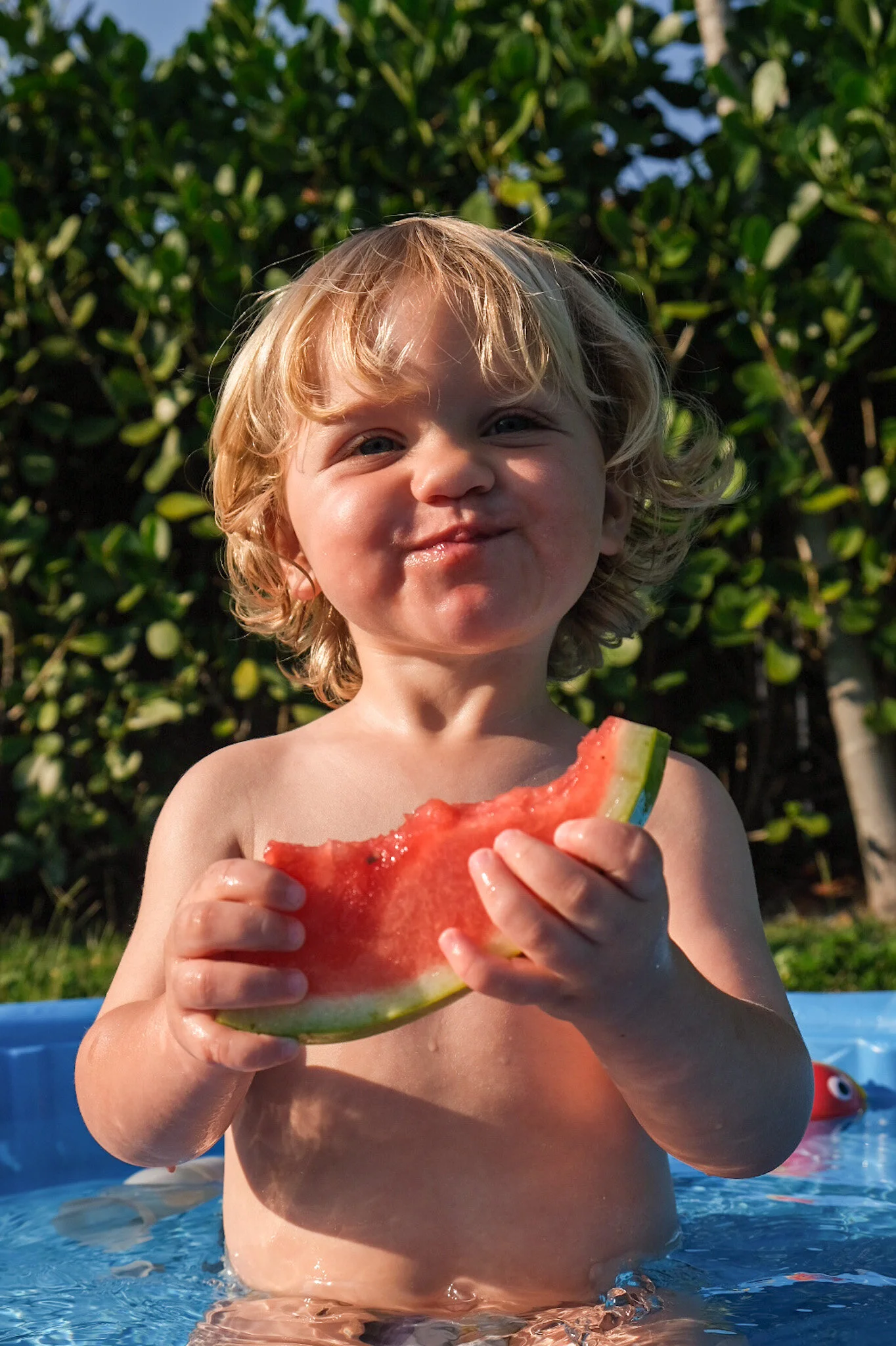 A young child with blonde curly hair smiling and holding a slice of watermelon while standing in a small pool outside, with green trees in the background.
