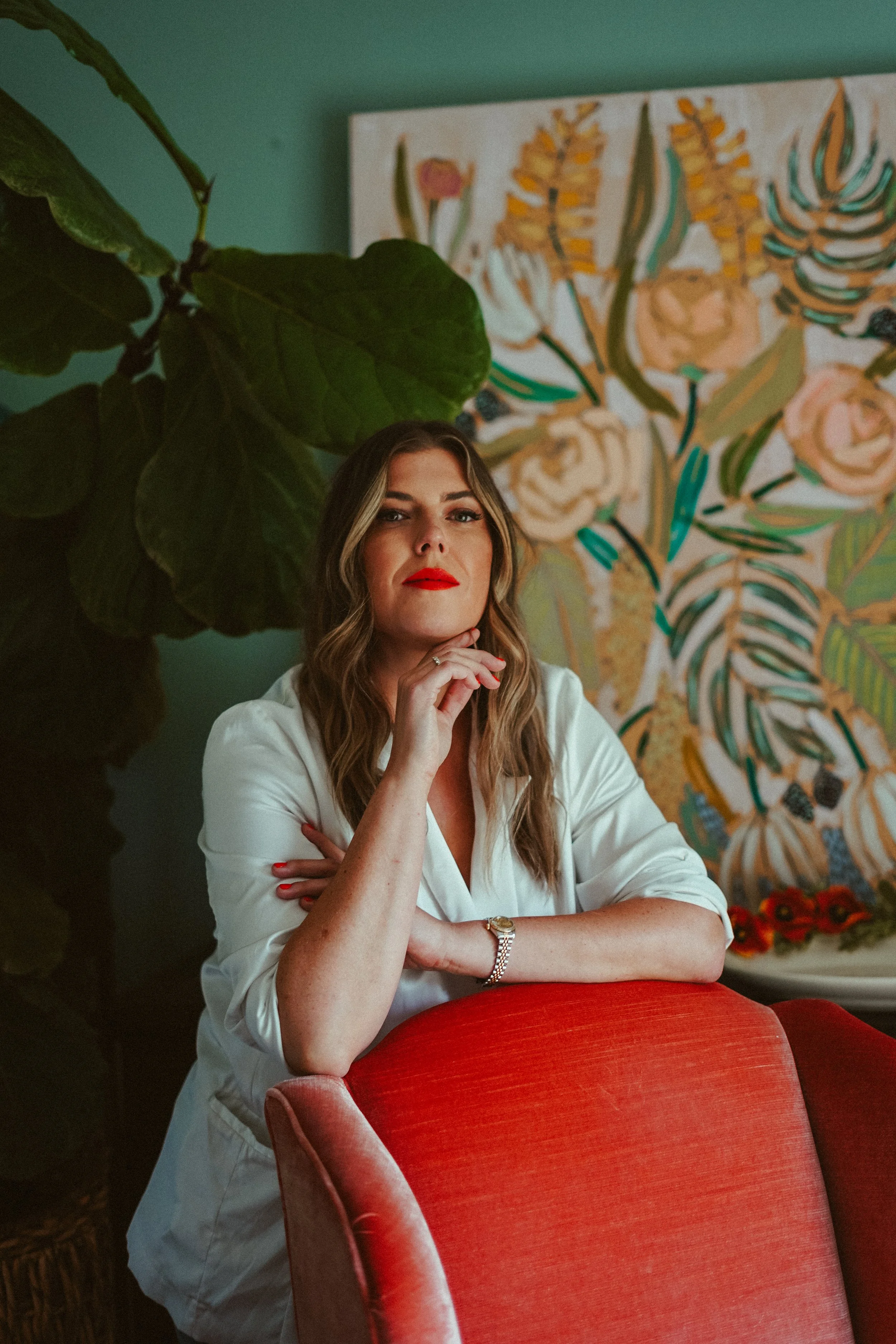 A woman with wavy blonde hair and red lipstick sitting on a red upholstered chair, resting her chin on her hand, in a room with large green leaves and a colorful floral painting in the background.