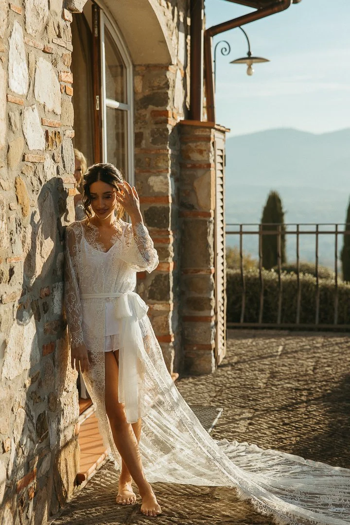 Woman in white lace robe standing barefoot outside on stone terrace, leaning against stone wall, smiling in sunlight with scenic mountain view in background.