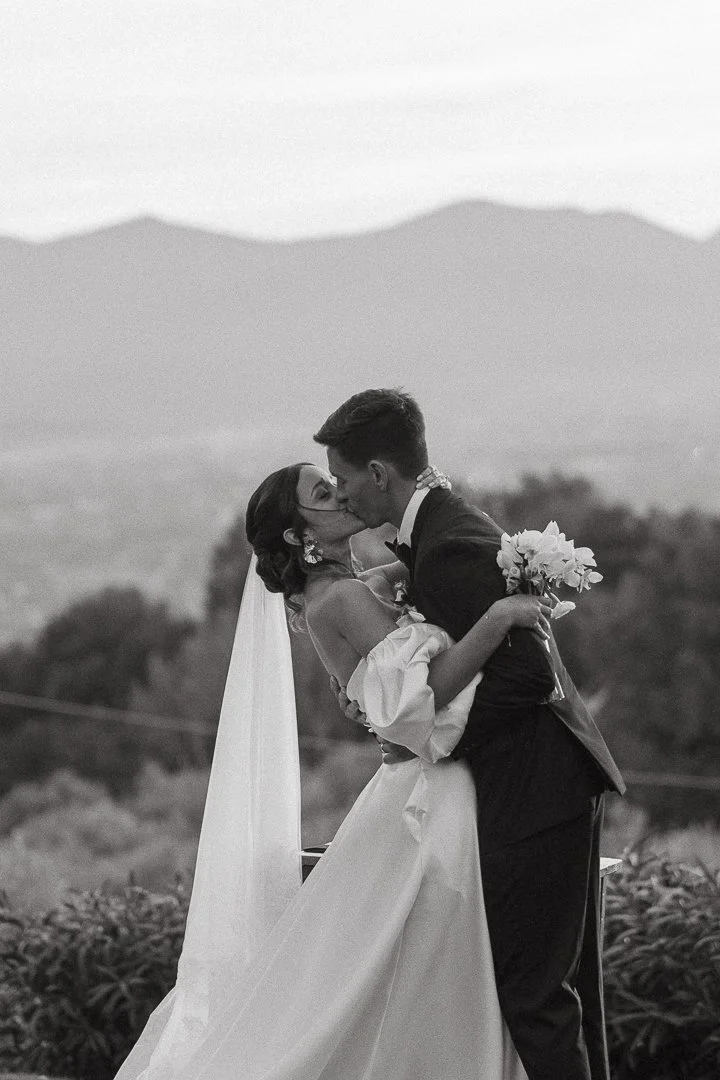A black-and-white photo of a bride and groom kissing outdoors, with mountains in the background. The bride is holding a bouquet, and they are dressed in wedding attire.
