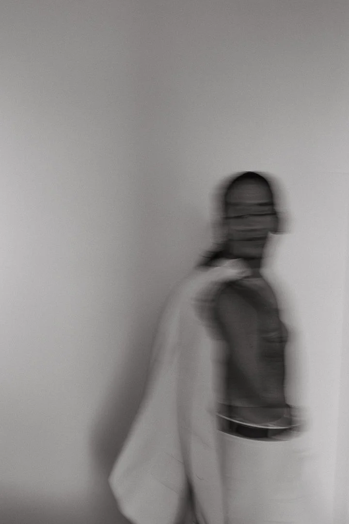 Blurry black and white photo of a woman with glasses standing against a plain wall, facing slightly to the right.