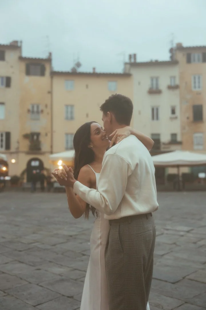 A young couple dancing and embracing outdoors in a European city square with old buildings in the background during the evening.