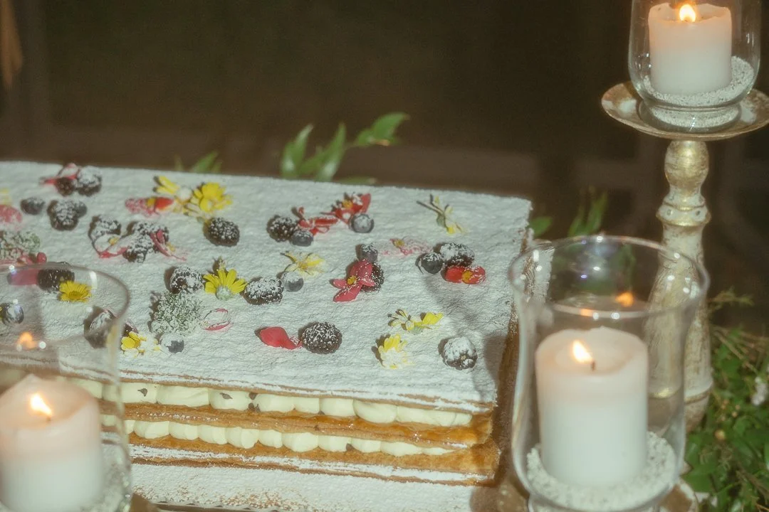 Layered cake with powdered sugar, blueberries, edible flowers, surrounded by lit candles in glass holders.
