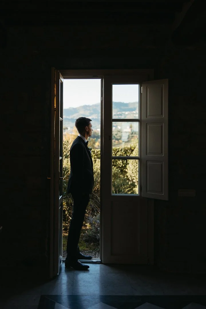 A man in a suit stands at an open door, looking outside at a scenic view of trees and mountains.