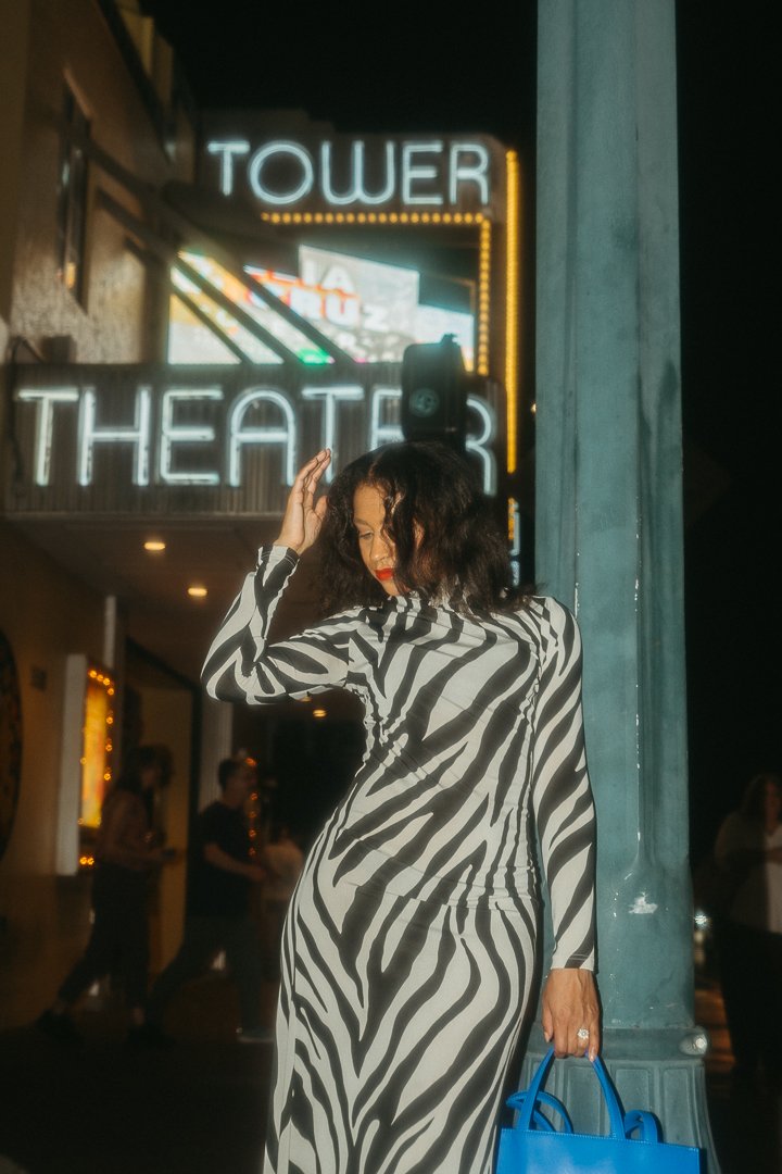 A woman in a zebra print dress standing outside a theater with neon signs at night, holding a blue handbag.