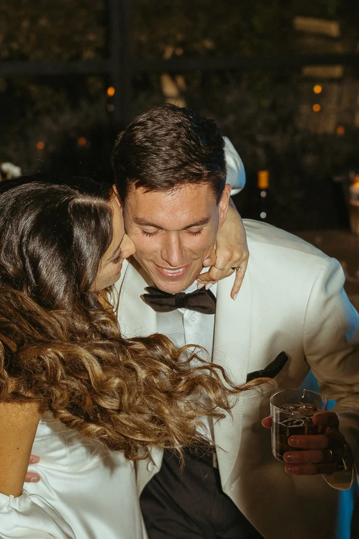A woman with long, curly brown hair hugging a man dressed in a white tuxedo with a black bow tie at night, holding a glass of dark beverage, with warm ambient lighting in the background.