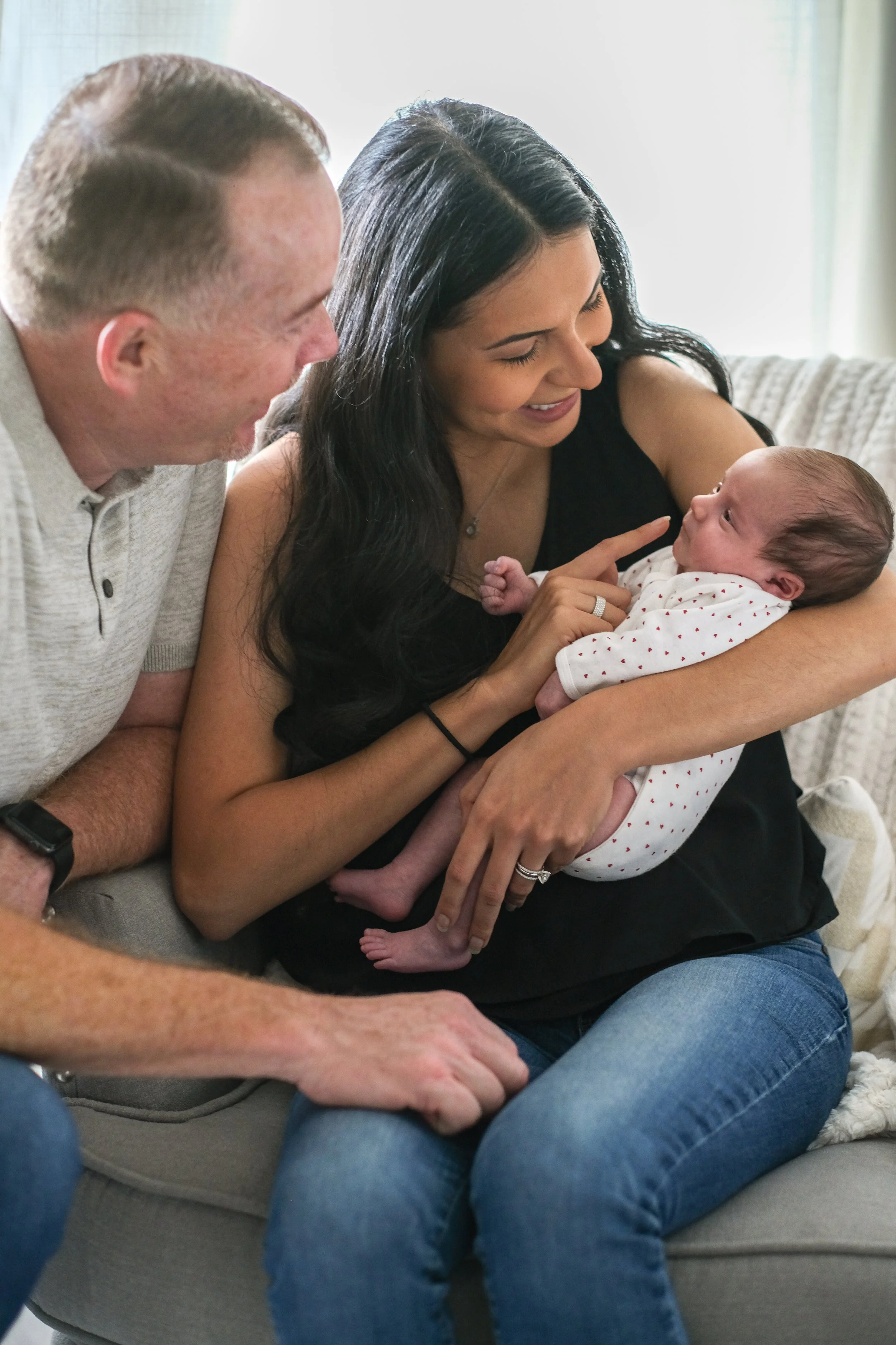 A woman holding a newborn baby on her lap, with an older man sitting nearby, all smiling and looking at each other.