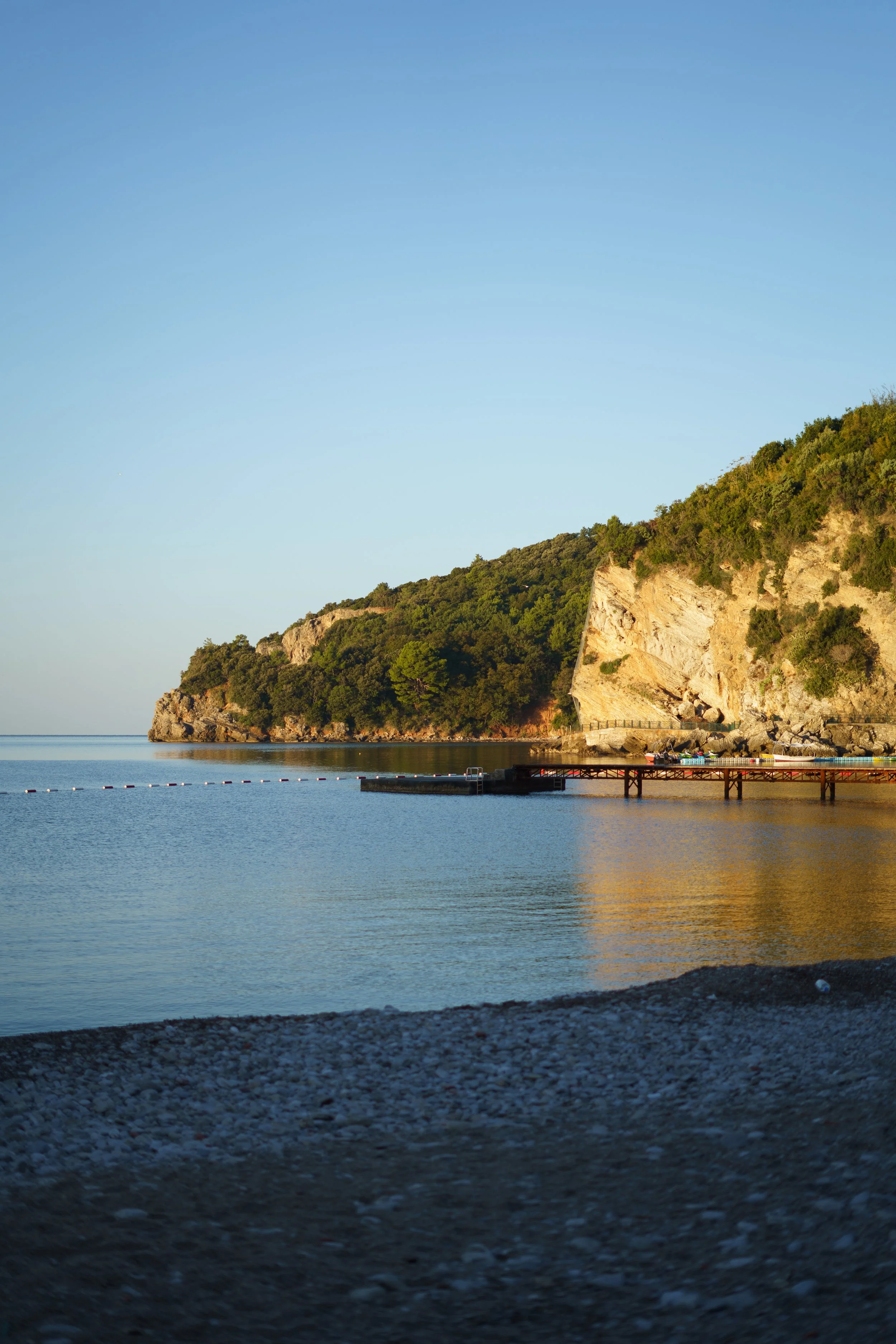A serene view of a beach with a pebble shoreline, calm water, and a hillside covered in trees, with cliffs on the right and a wooden pier extending into the water under a clear blue sky.