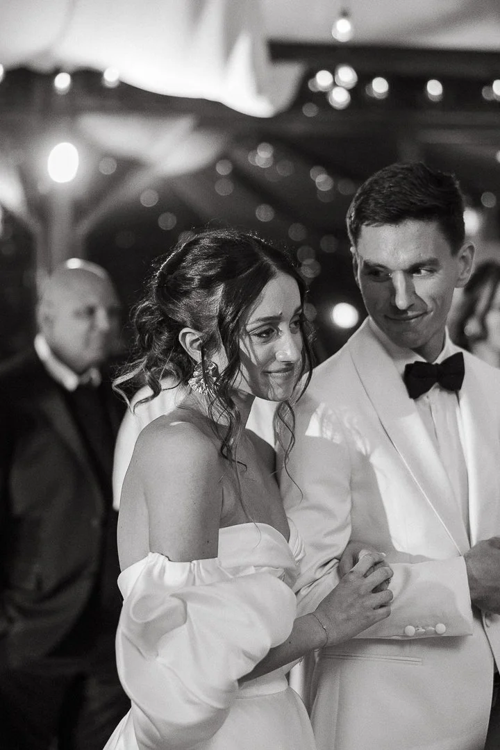 Black and white photo of a bride and groom at a wedding reception, with the bride in an off-the-shoulder dress and the groom in a tuxedo, standing close together in a decorated venue.