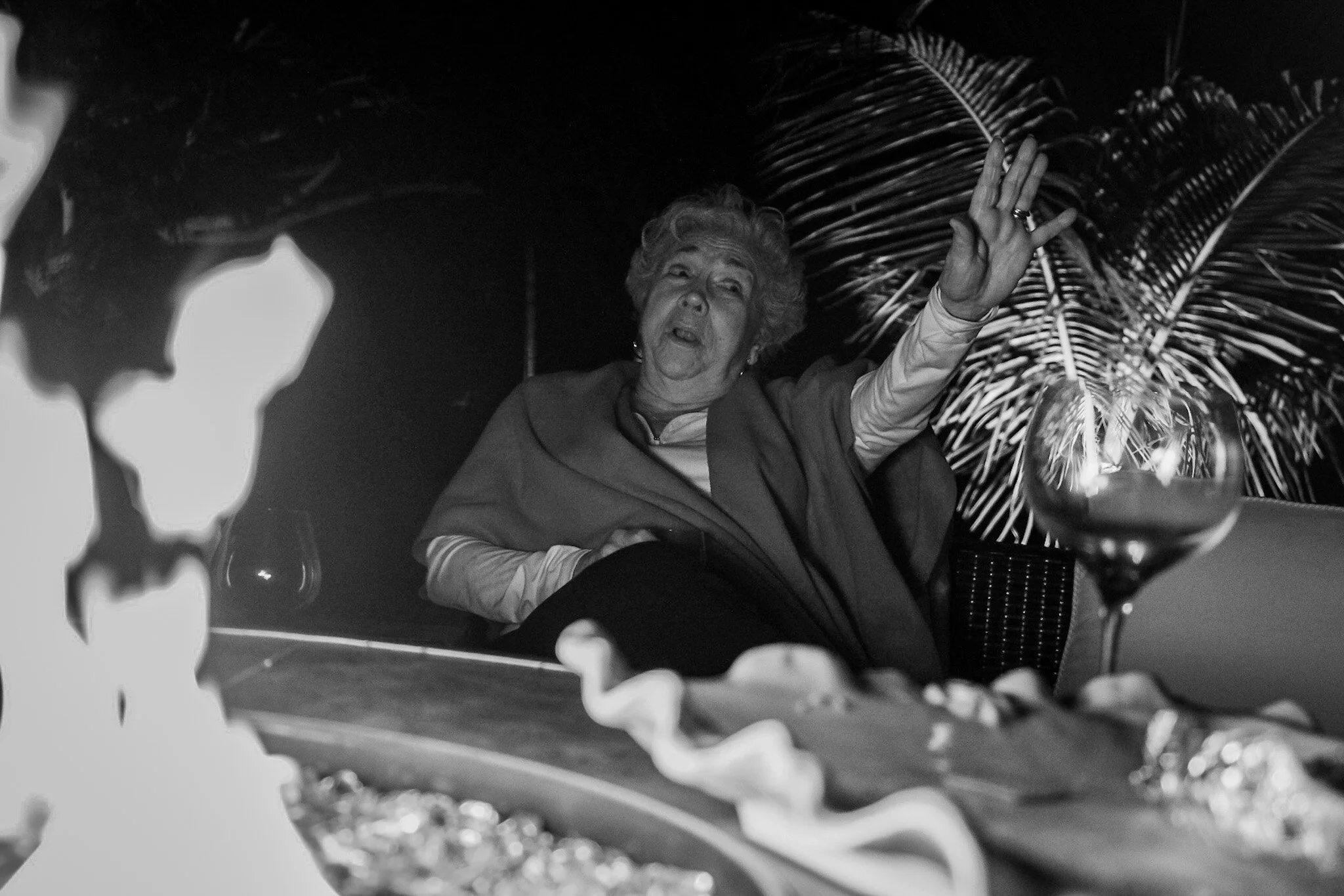 Older woman with curly hair gesturing with her right hand during social gathering, with palm leaves in the background and a glass of drink on the table in front of her.