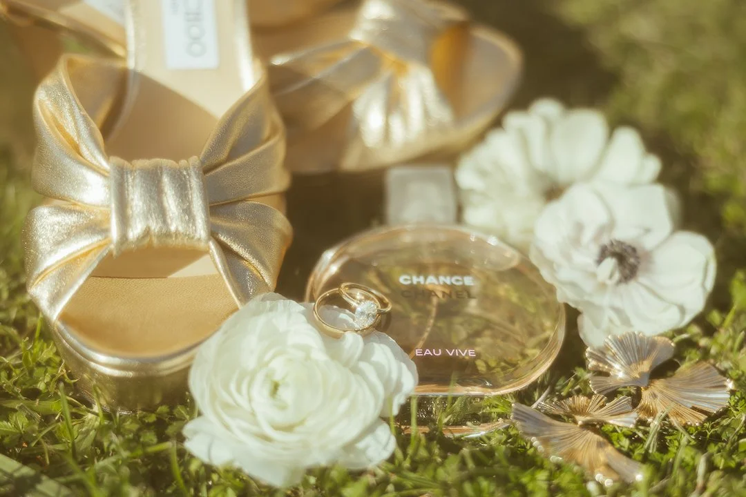 Gold gift box with bow, Chanel Chance Eau Vive perfume bottle, white flowers, gold decorative elements, and a ring outdoors on grass.