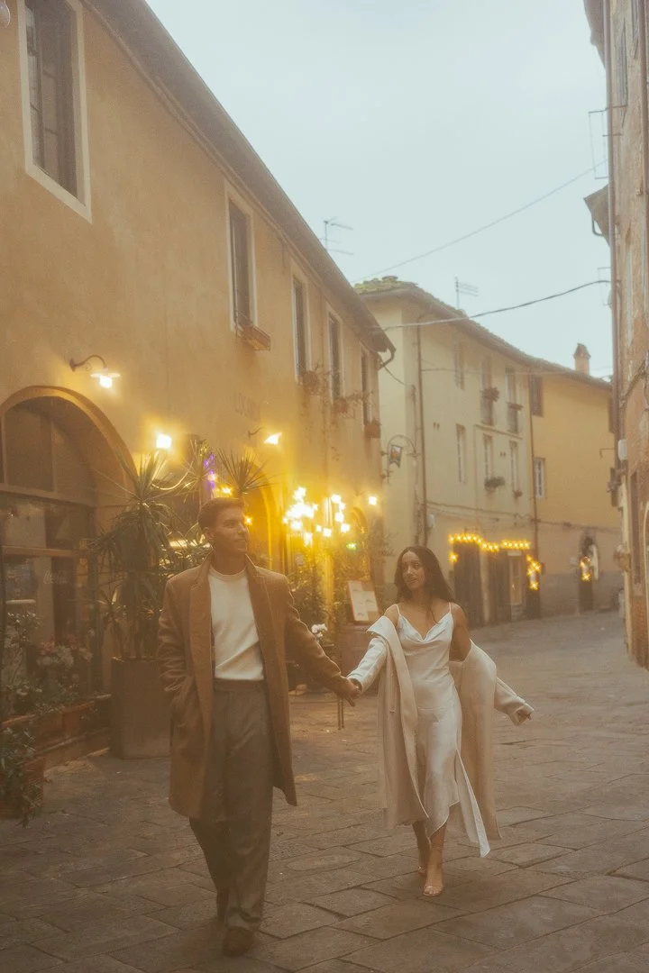 A couple walking hand in hand on a cobblestone street in a European city at dusk, with warm street lighting and outdoor cafes.