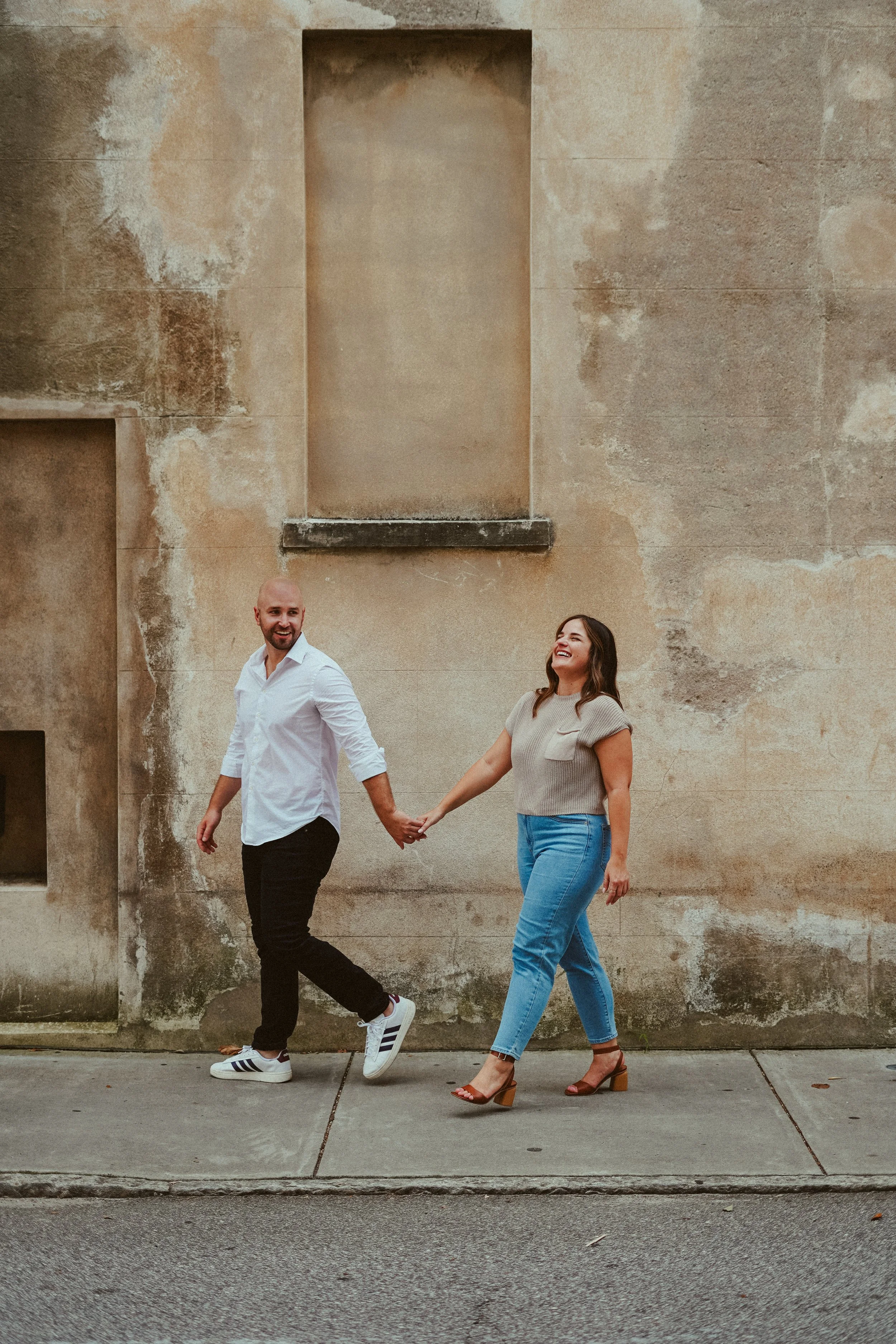 A smiling man and woman holding hands and walking on the sidewalk in front of an aged, textured beige wall.