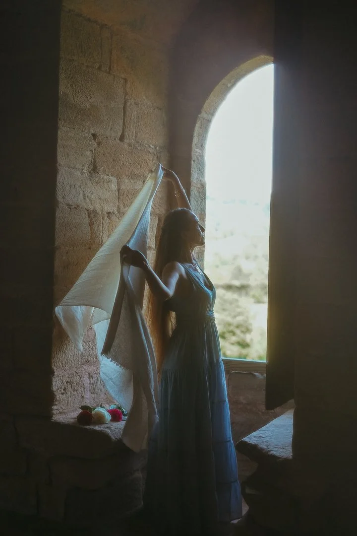 A woman in a long dress stands by an arched window, holding a piece of fabric above her head and gazing outside, with sunlight illuminating her silhouette inside a stone structure.
