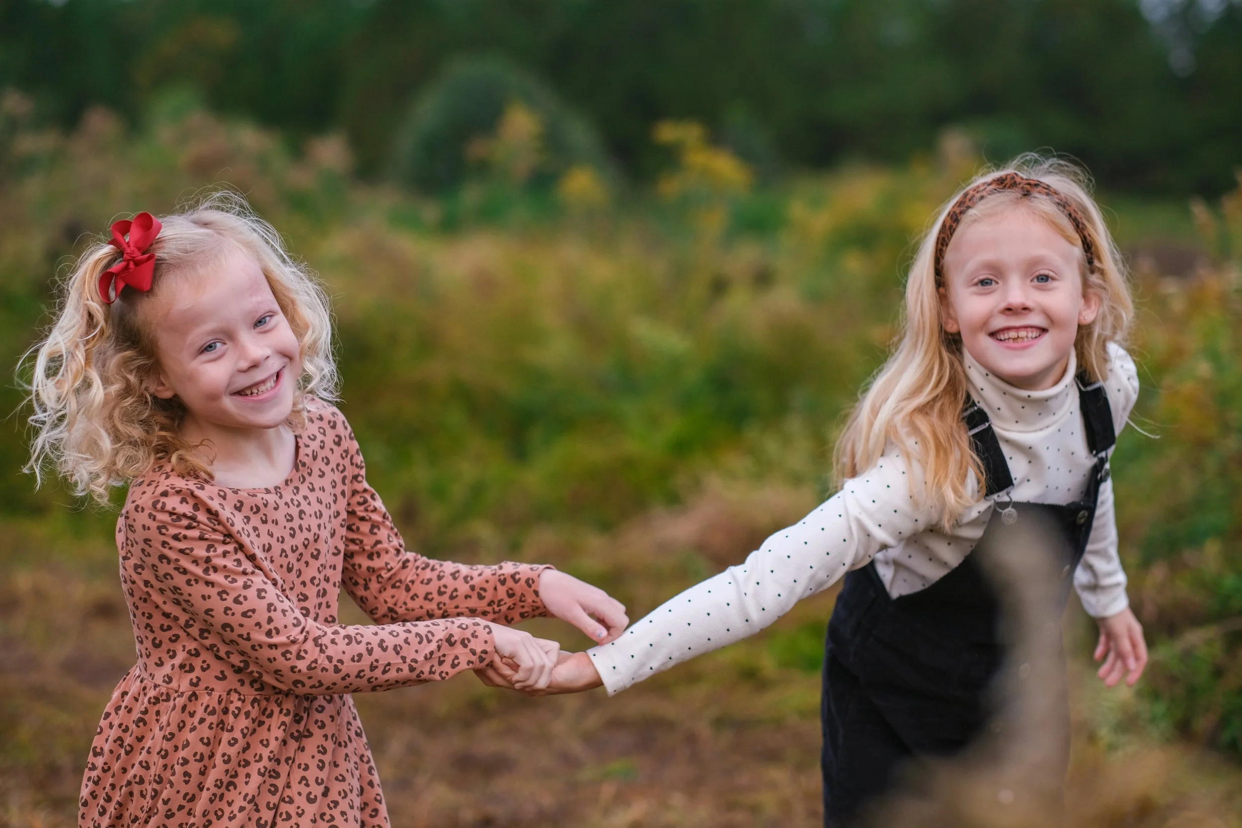 Two young girls smiling and holding hands outdoors in a grassy area with trees in the background.