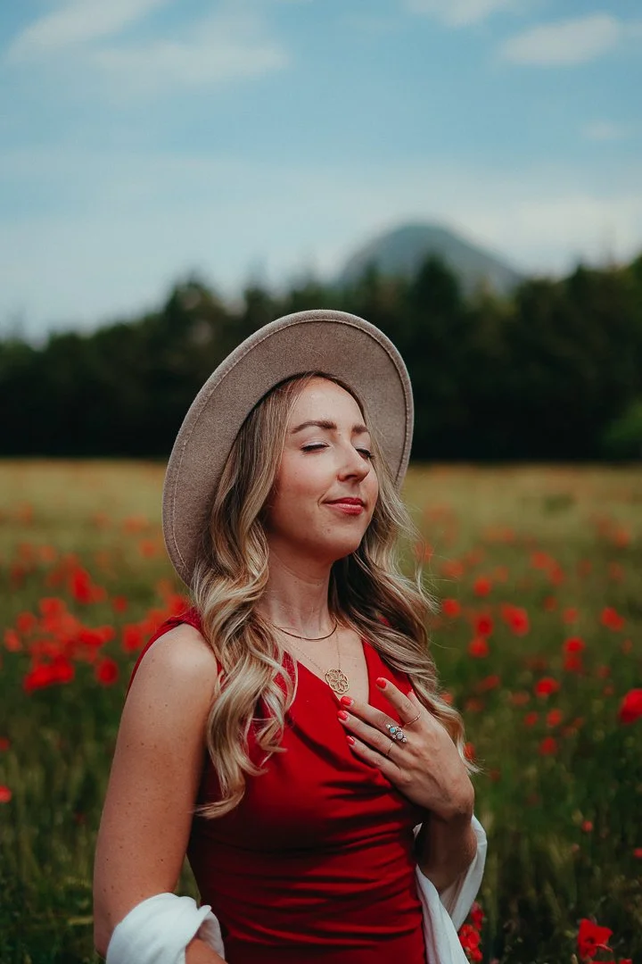 A woman with long blonde hair wearing a beige hat, red dress, and white jacket draped over her arms stands in a field of red flowers, with trees and a mountain in the background.