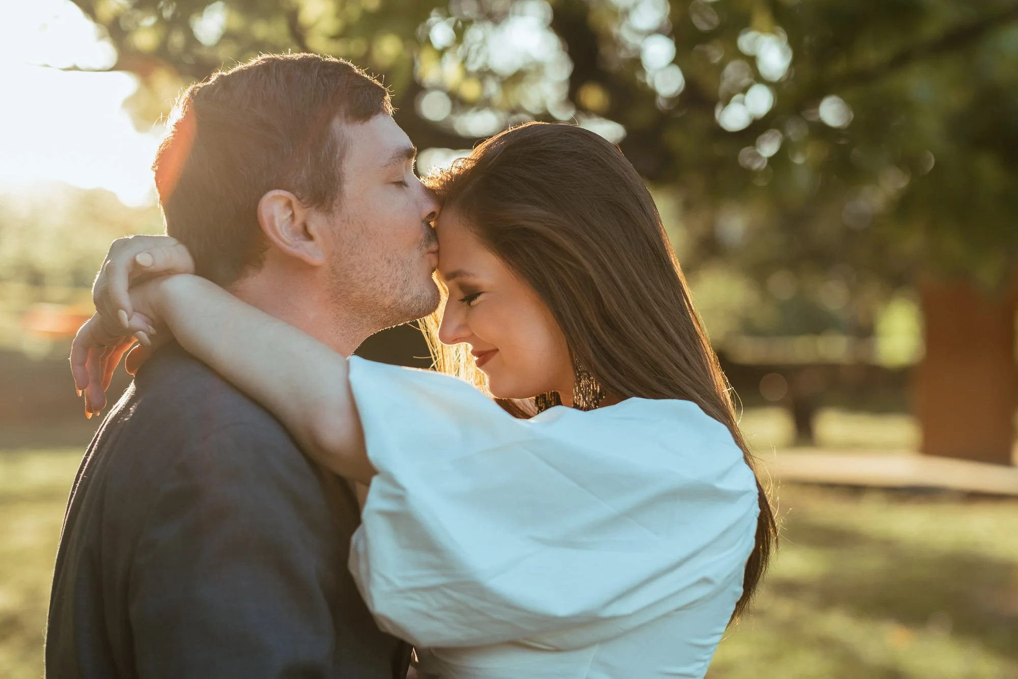 A couple sharing an intimate moment outdoors during sunset, with the man kissing the woman on the forehead and the woman smiling with her eyes closed, surrounded by trees and soft sunlight.