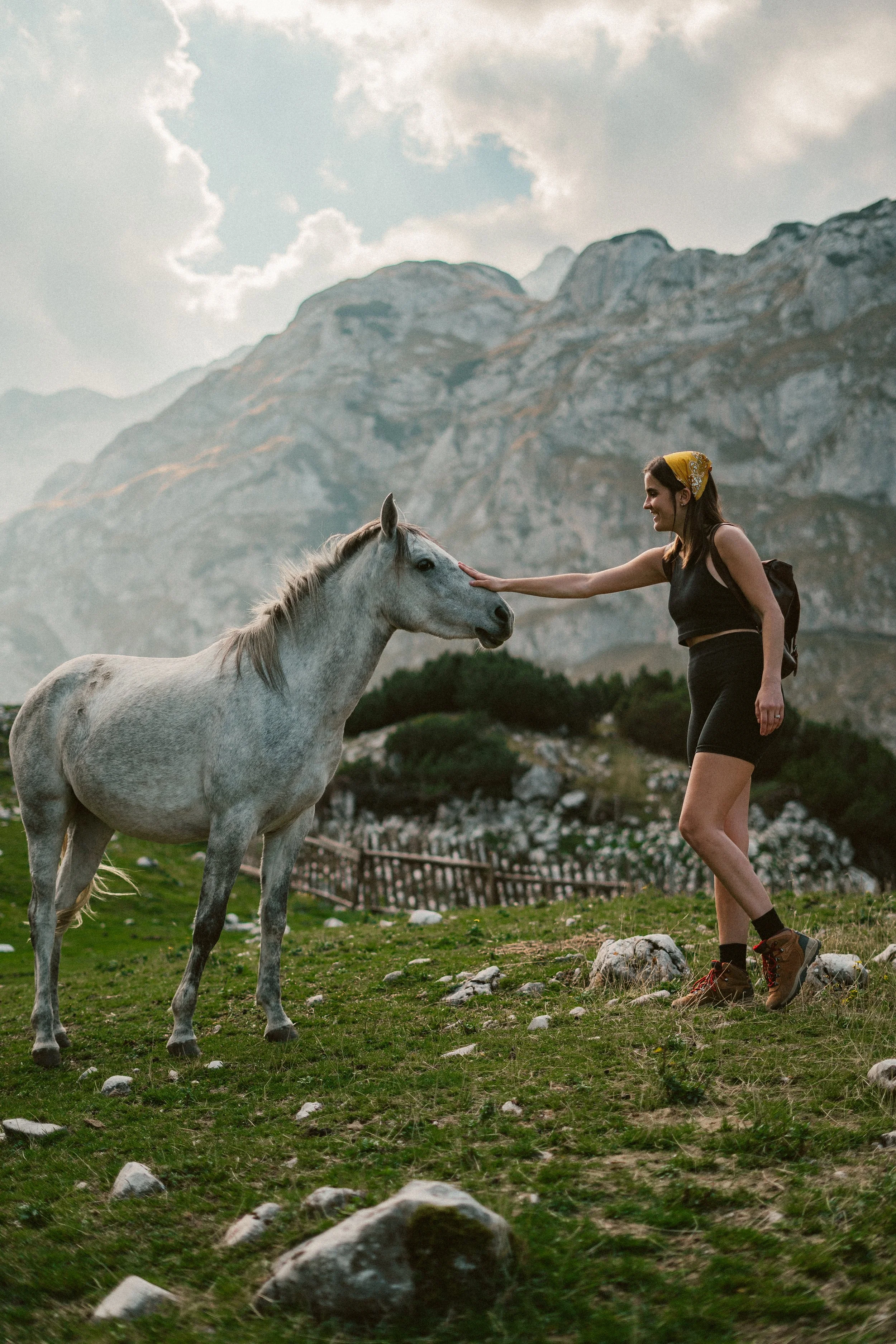 A woman with a yellow headscarf touching a gray horse's face in a mountainous landscape.