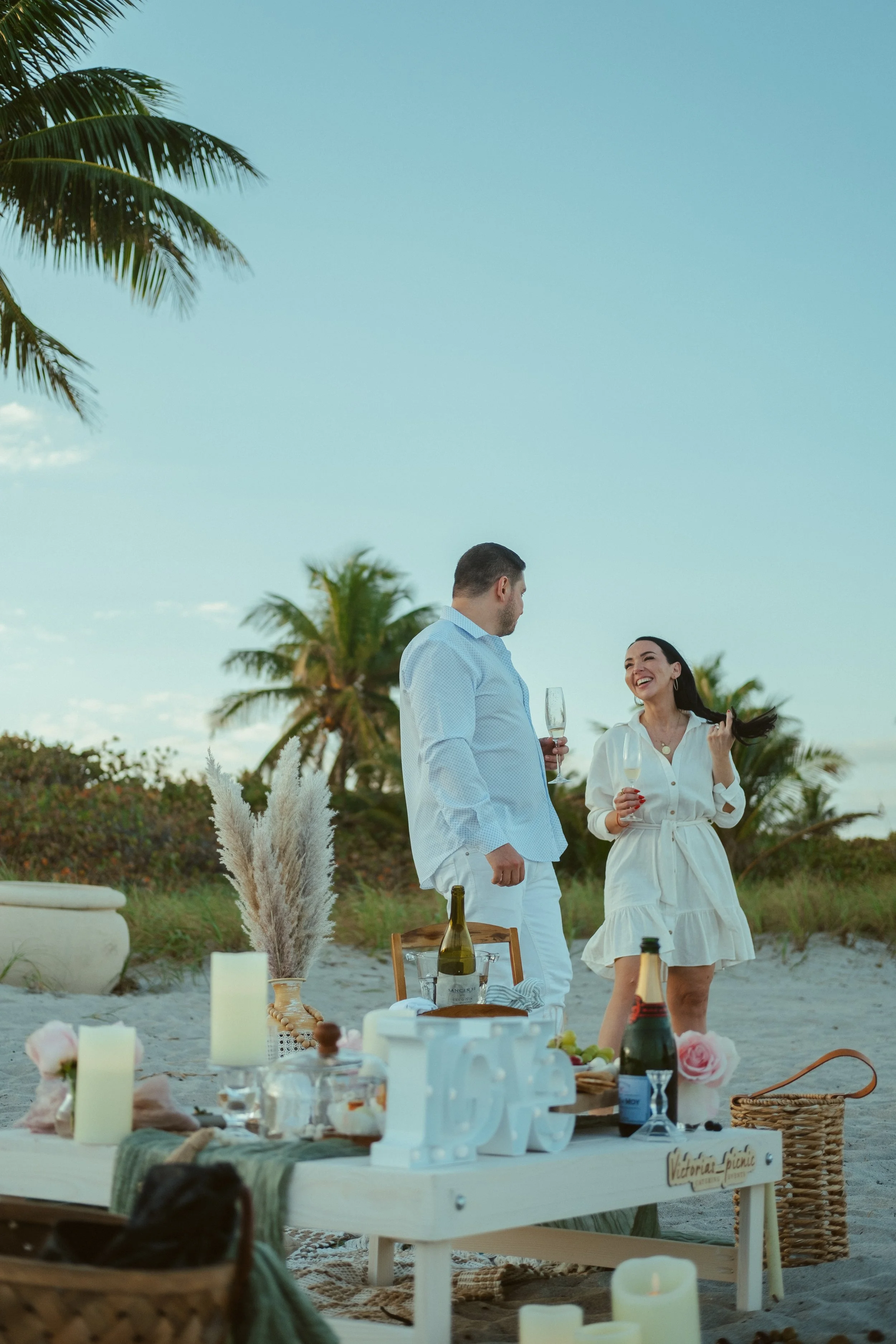 A couple enjoys a romantic picnic on the beach, with palm trees, candles, and a spread of food and drinks around a low table, during the evening.